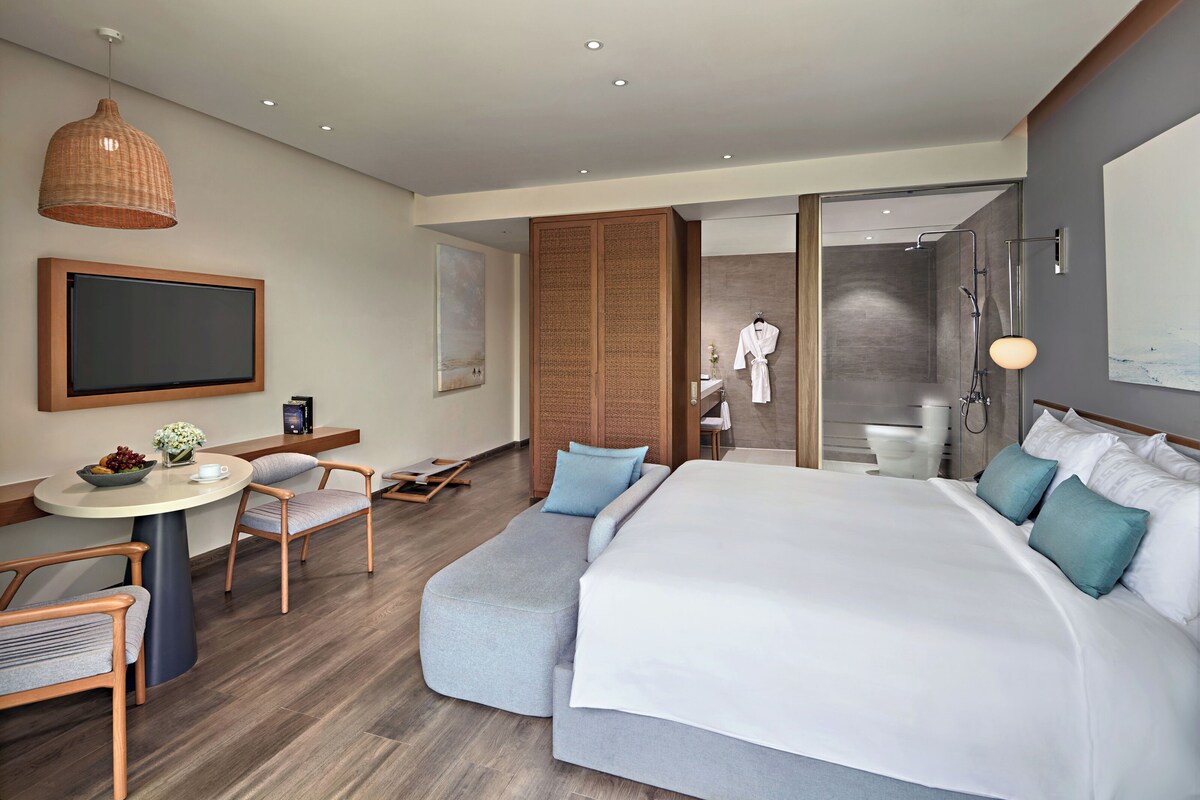 Una habitación de hotel moderna con una cama grande, una zona de estar con sillas, una mesita con un frutero, un televisor y una zona de ducha.