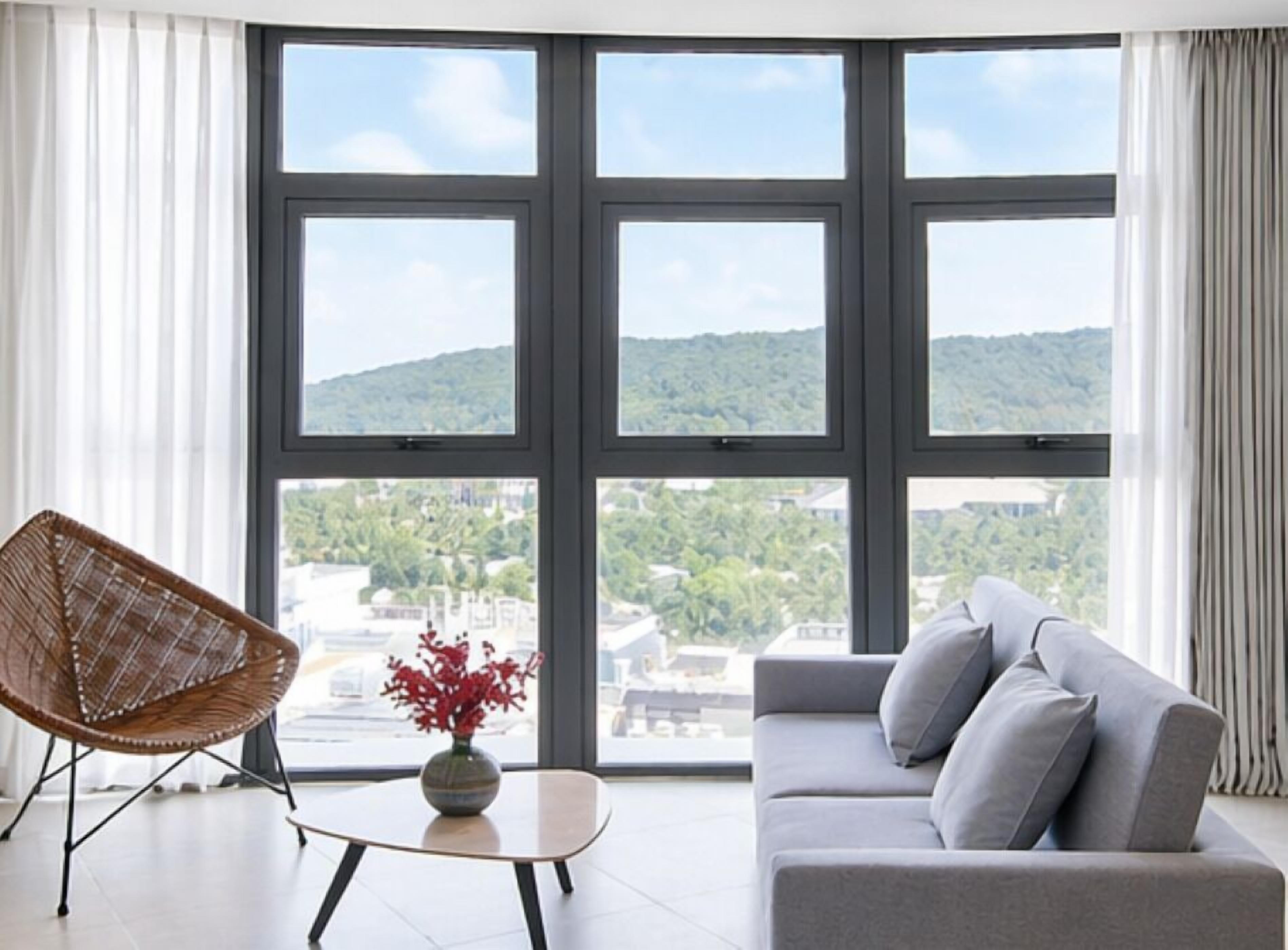 A modern living room with a large window, a sofa, a wicker chair, and a small table with a vase.