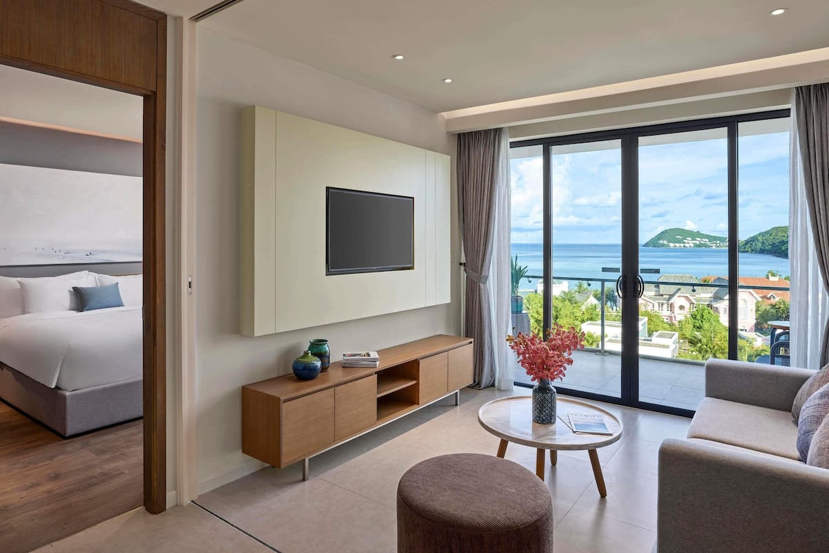 Habitación de hotel con cama, televisor montado en la pared, sofá, mesa de centro redonda y vista al mar.
