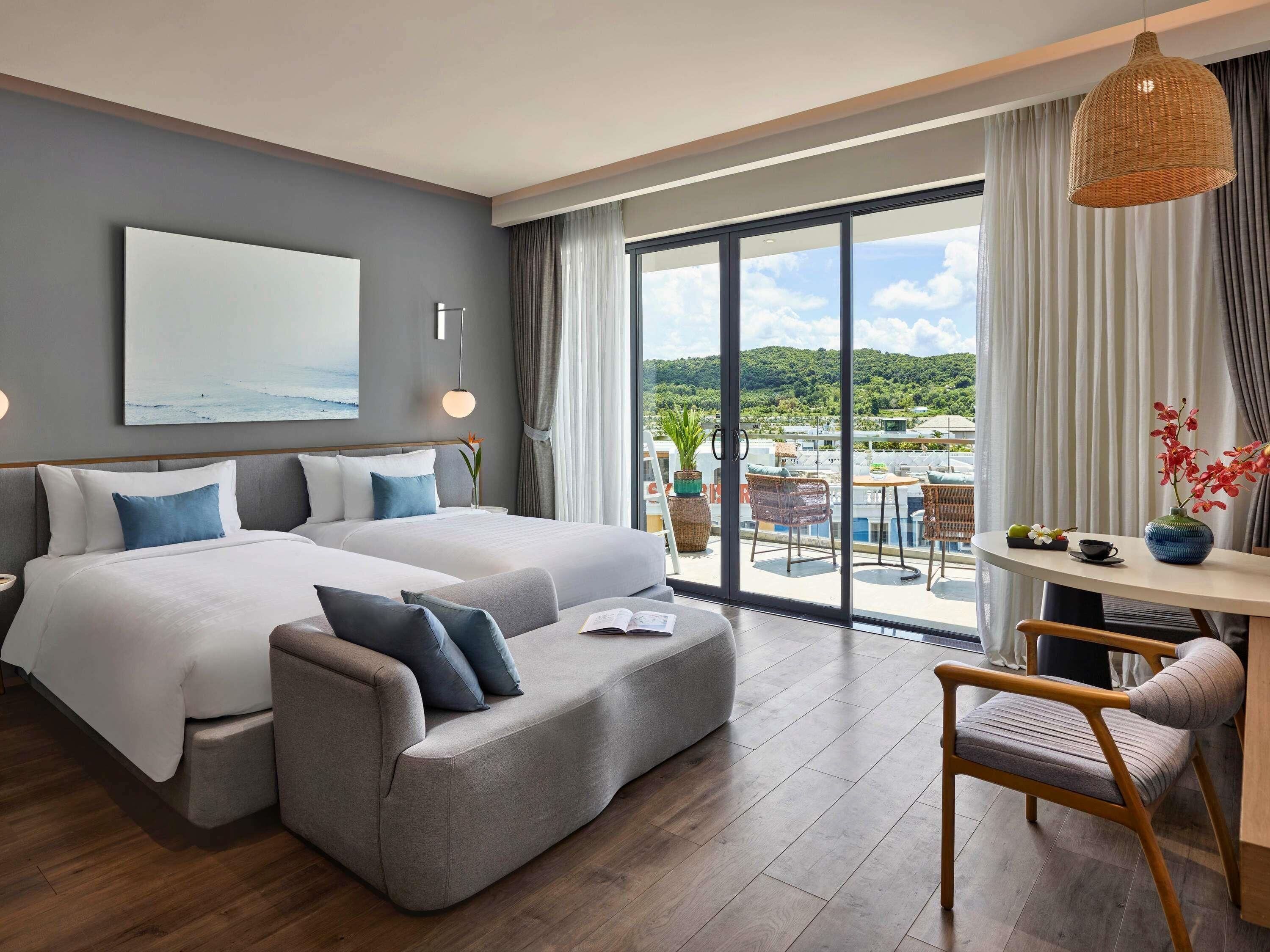 A modern hotel room with a large bed, a sofa, a dining table, and a view of the landscape.