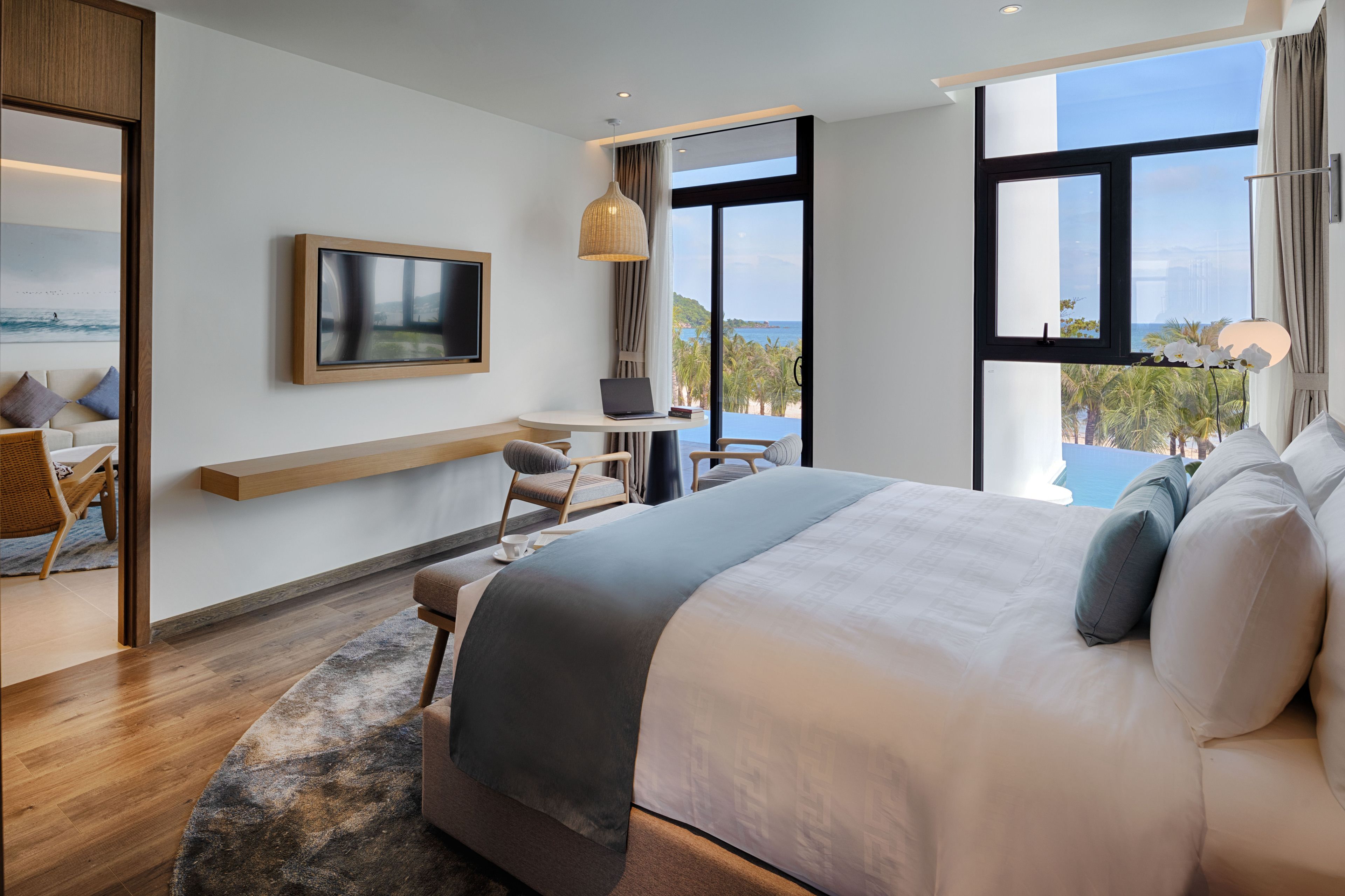 A modern hotel room with a large bed, a desk with a laptop, a TV, and a view of the sea.