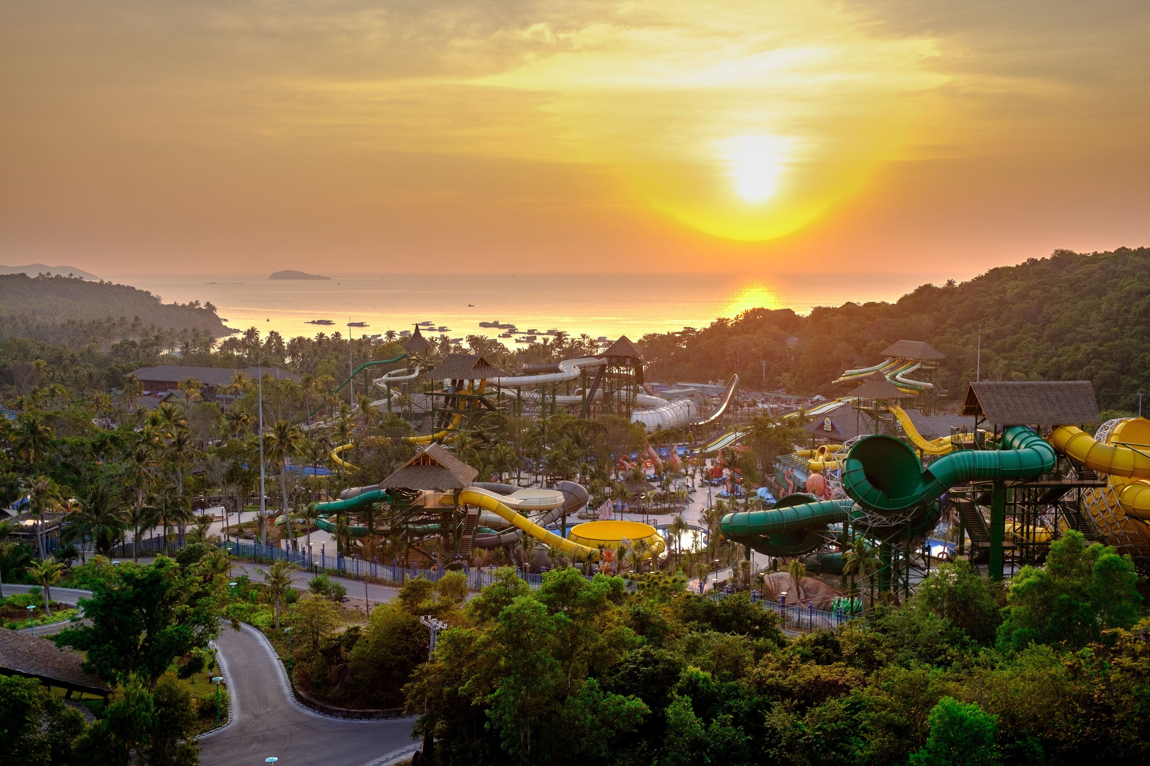 Un parc aquatique doté de nombreux toboggans et de plusieurs piscines, entouré d’une végétation luxuriante, avec un coucher de soleil en toile de fond.