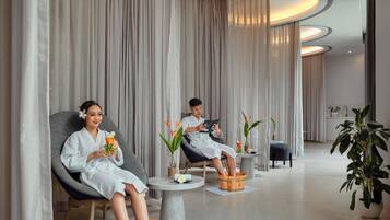 A spa room with two people relaxing in chairs, a wooden foot bath, and a small table with a drink and flowers.