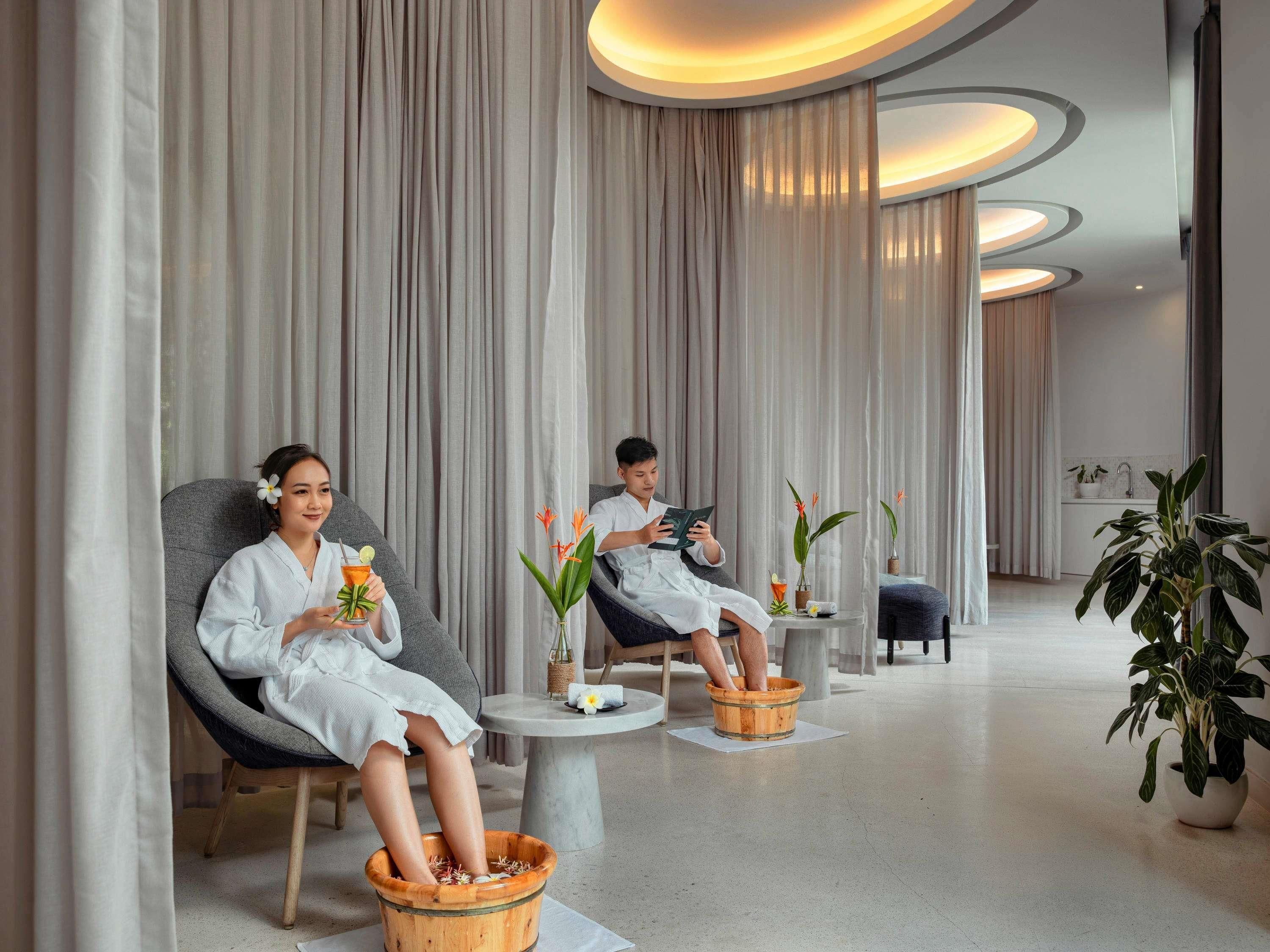 A spa room with two people relaxing in chairs, a wooden foot bath, and a small table with a drink and flowers.
