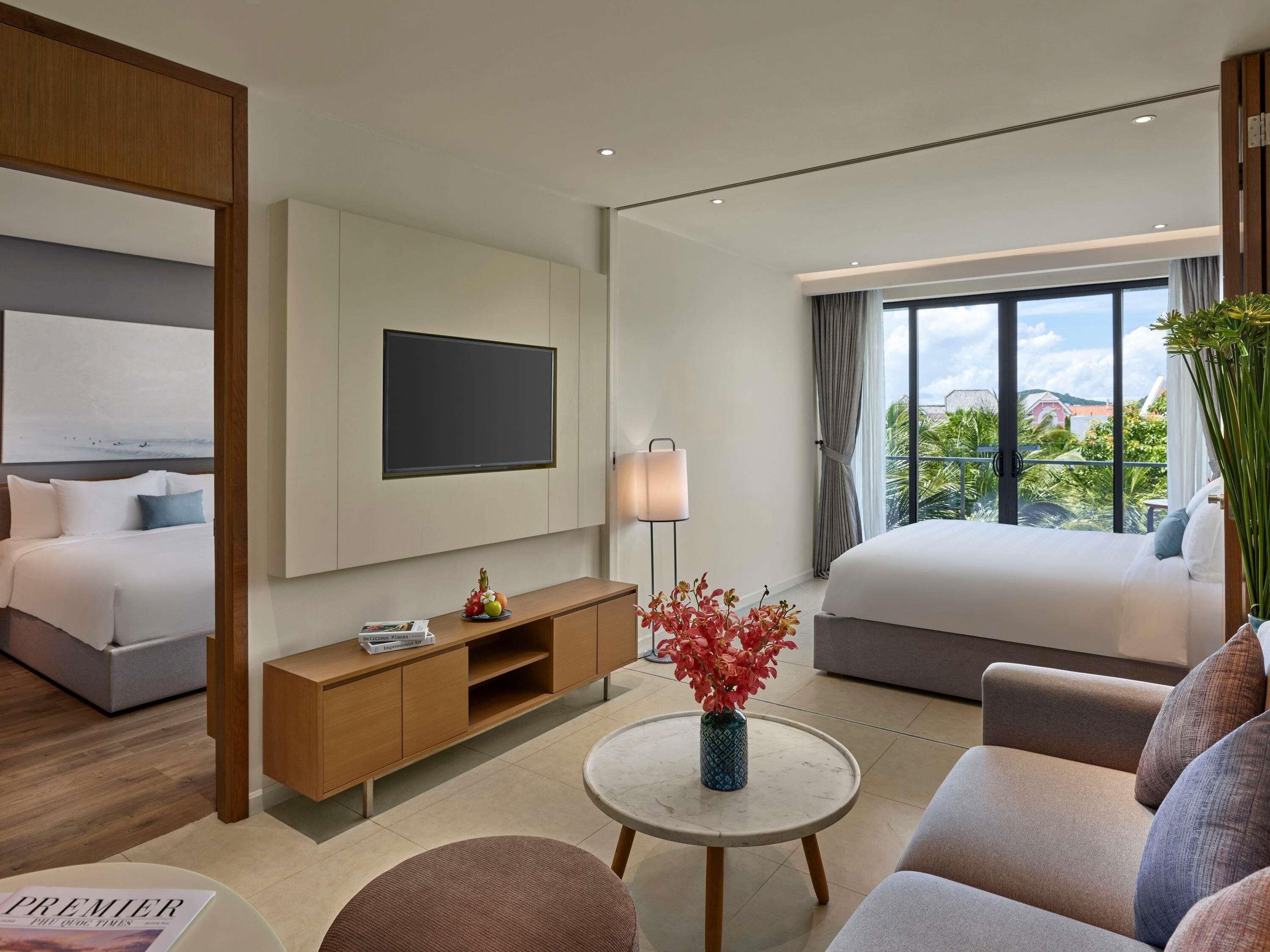 A modern hotel room with a bed, a TV, a bedside table, a sofa, and a view of the outdoors.