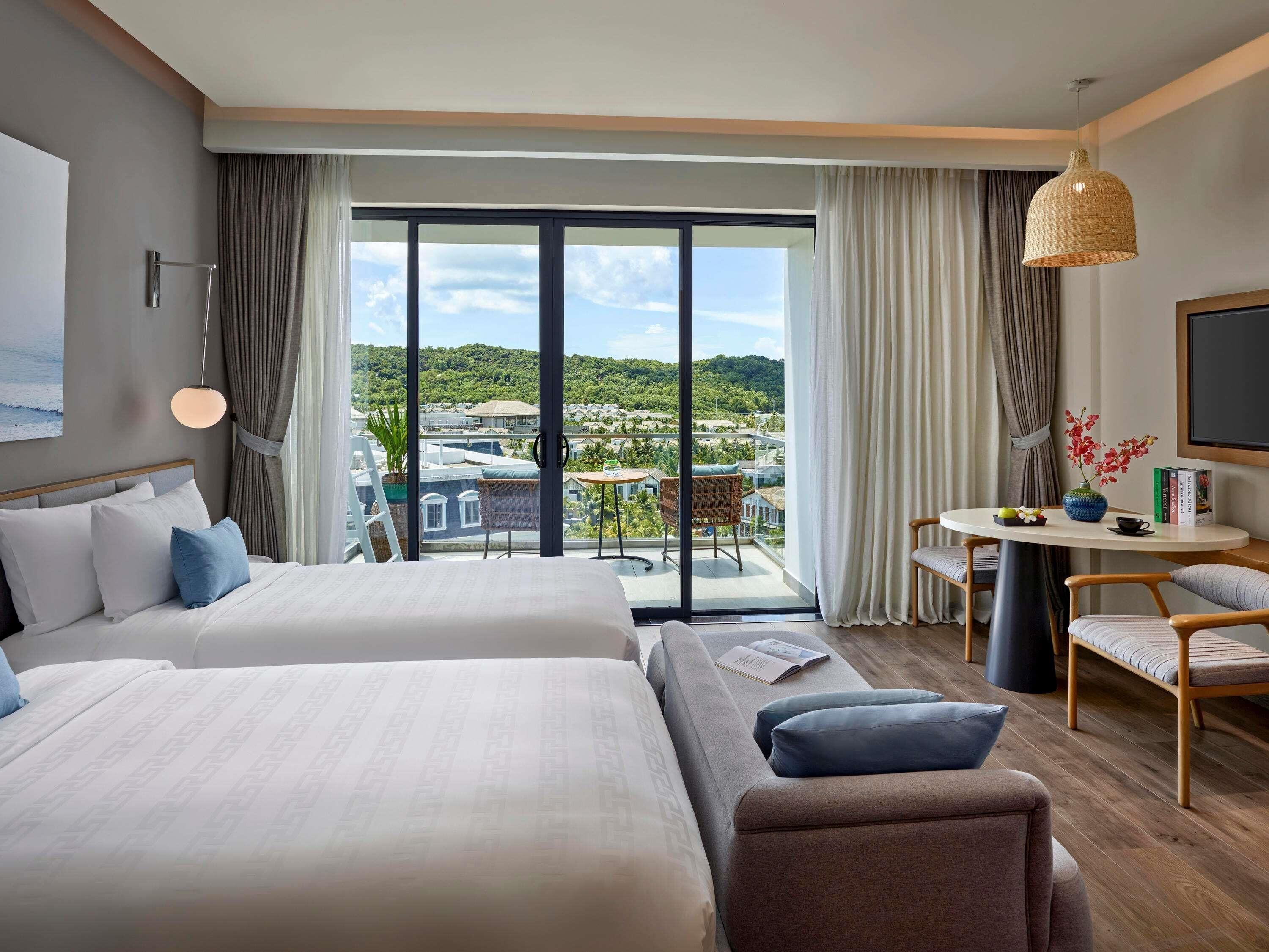 A hotel room with two beds, a balcony with a view, a small table with a vase, and a TV on the wall.