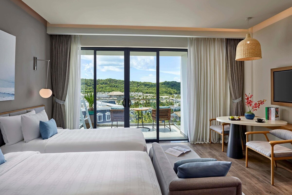 Habitación de hotel con dos camas, un balcón con vistas, una mesita con un jarrón y un televisor en la pared.