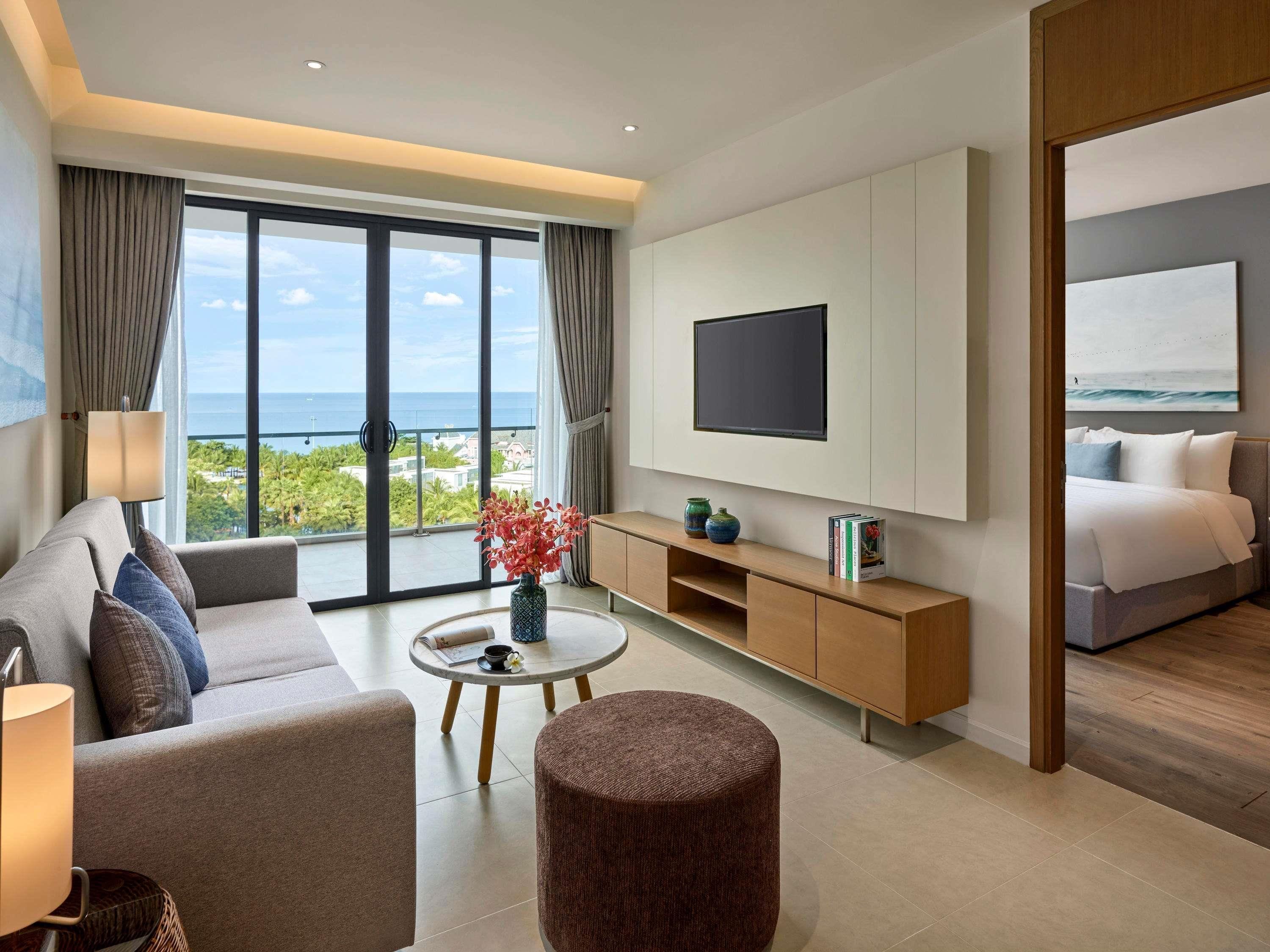 A modern hotel room with a large window, a flat-screen TV, a sofa, a coffee table, and a bed.