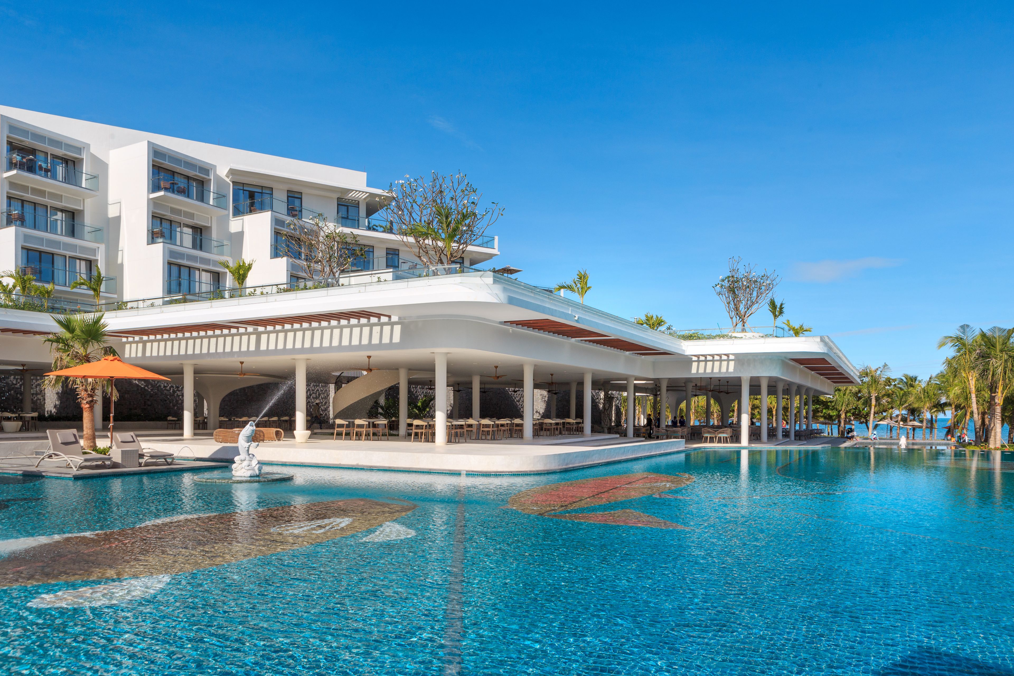 A modern hotel with a large pool, outdoor seating, and palm trees.