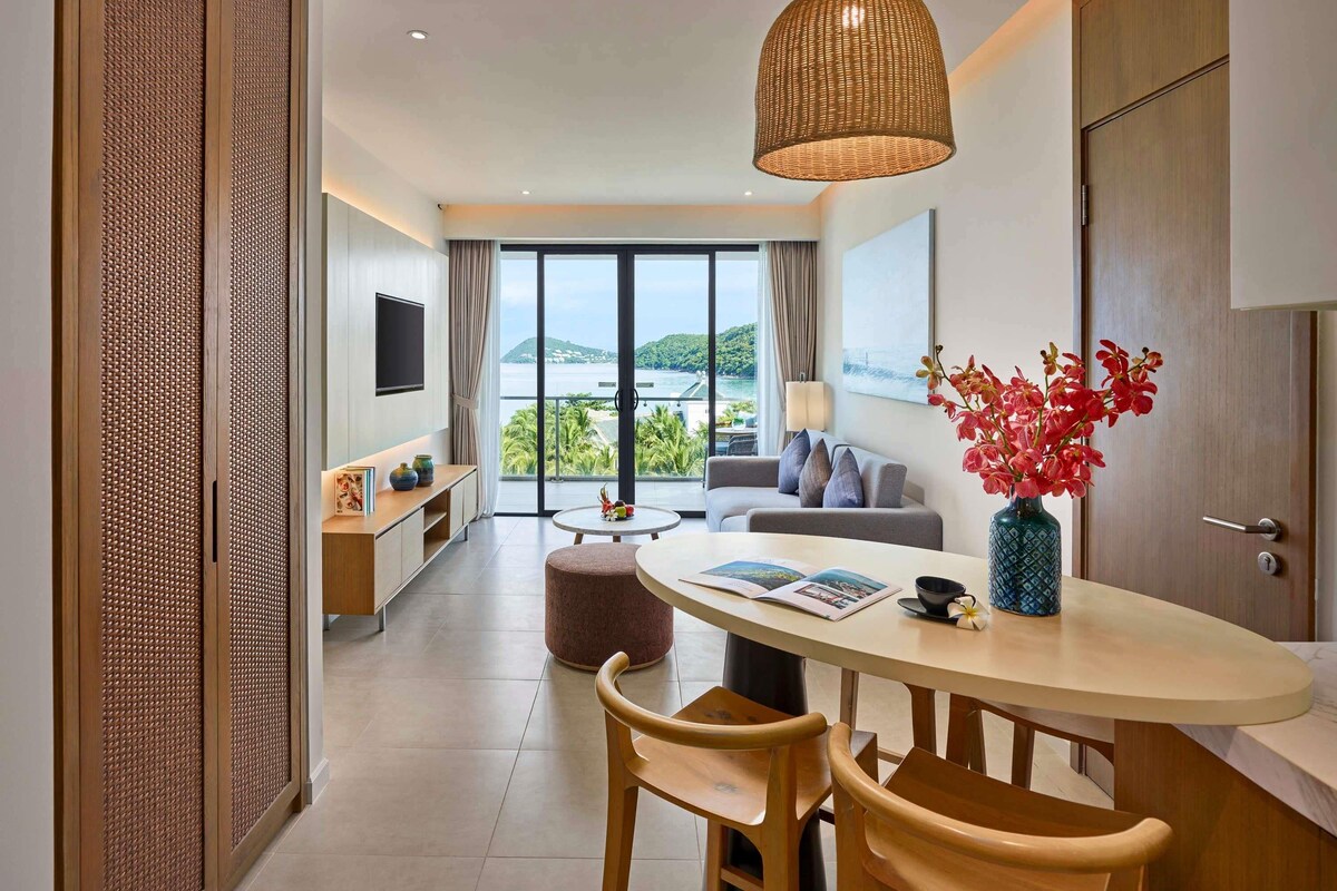 A modern hotel room with a dining area, a sofa, a TV, and a view of the sea.