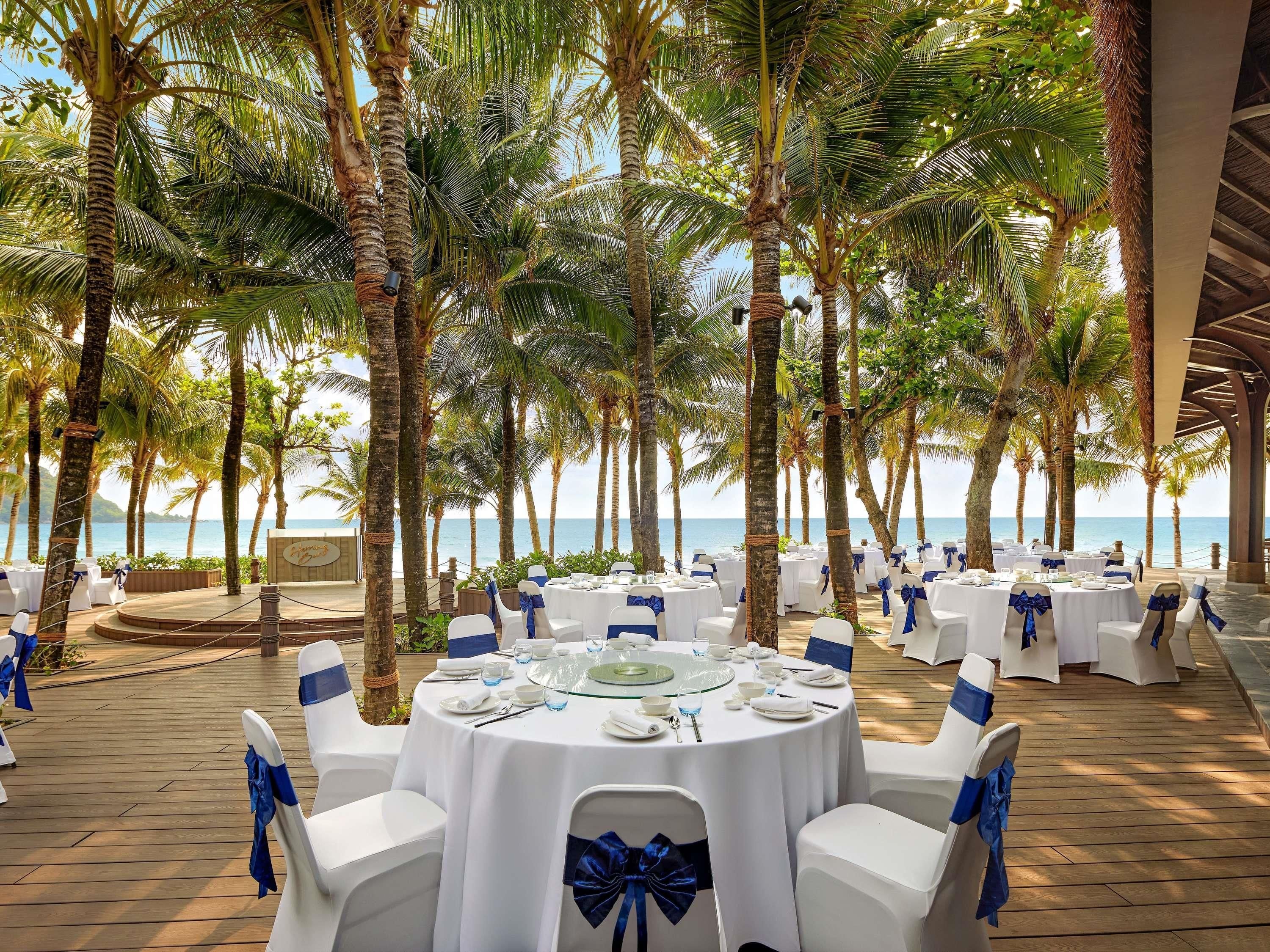 An outdoor dining area with round tables, white chairs, and blue sashes, surrounded by palm trees and overlooking a body of water.