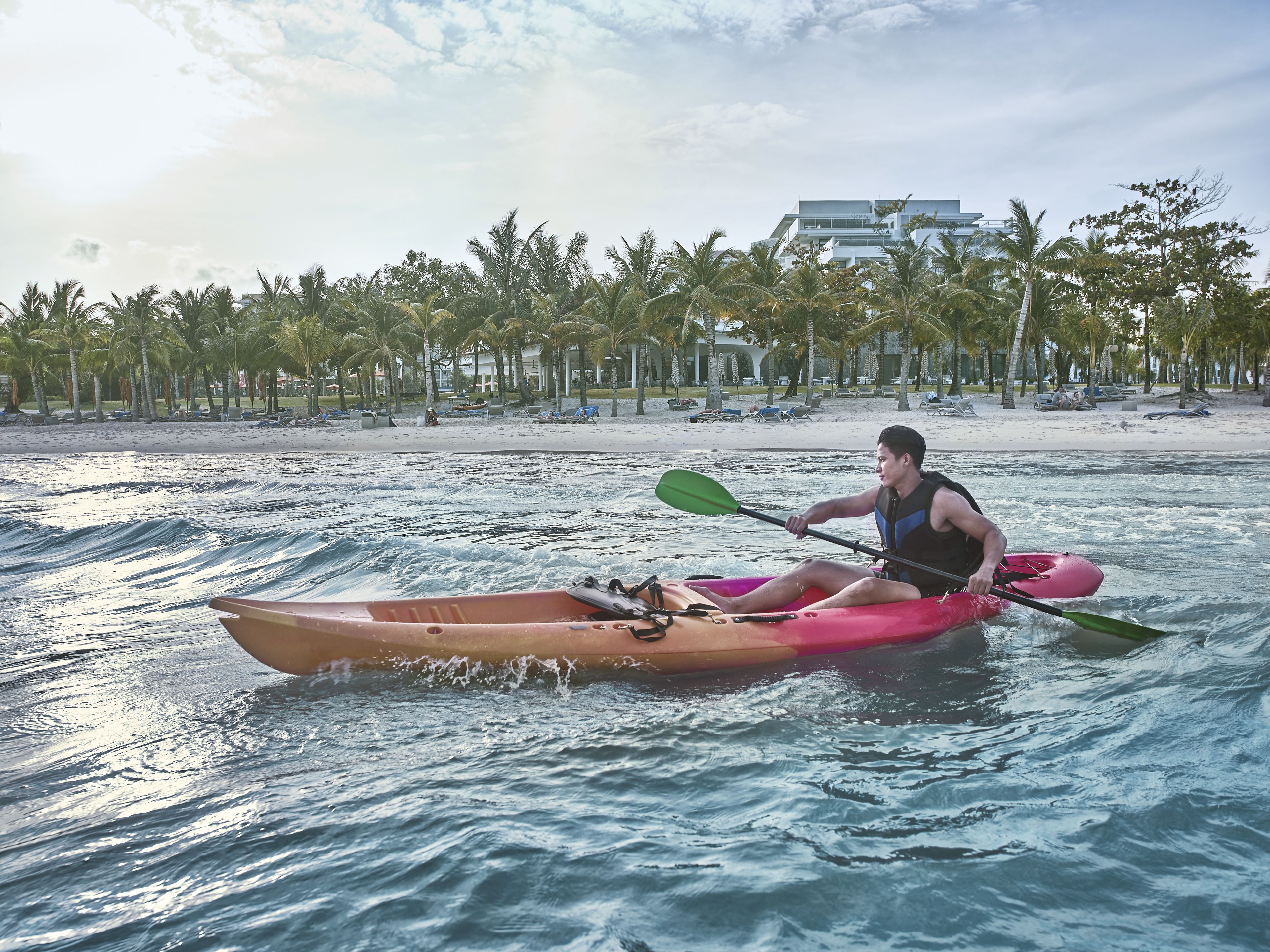 Una persona en kayak sobre un mar tranquilo, con una playa y palmeras al fondo.