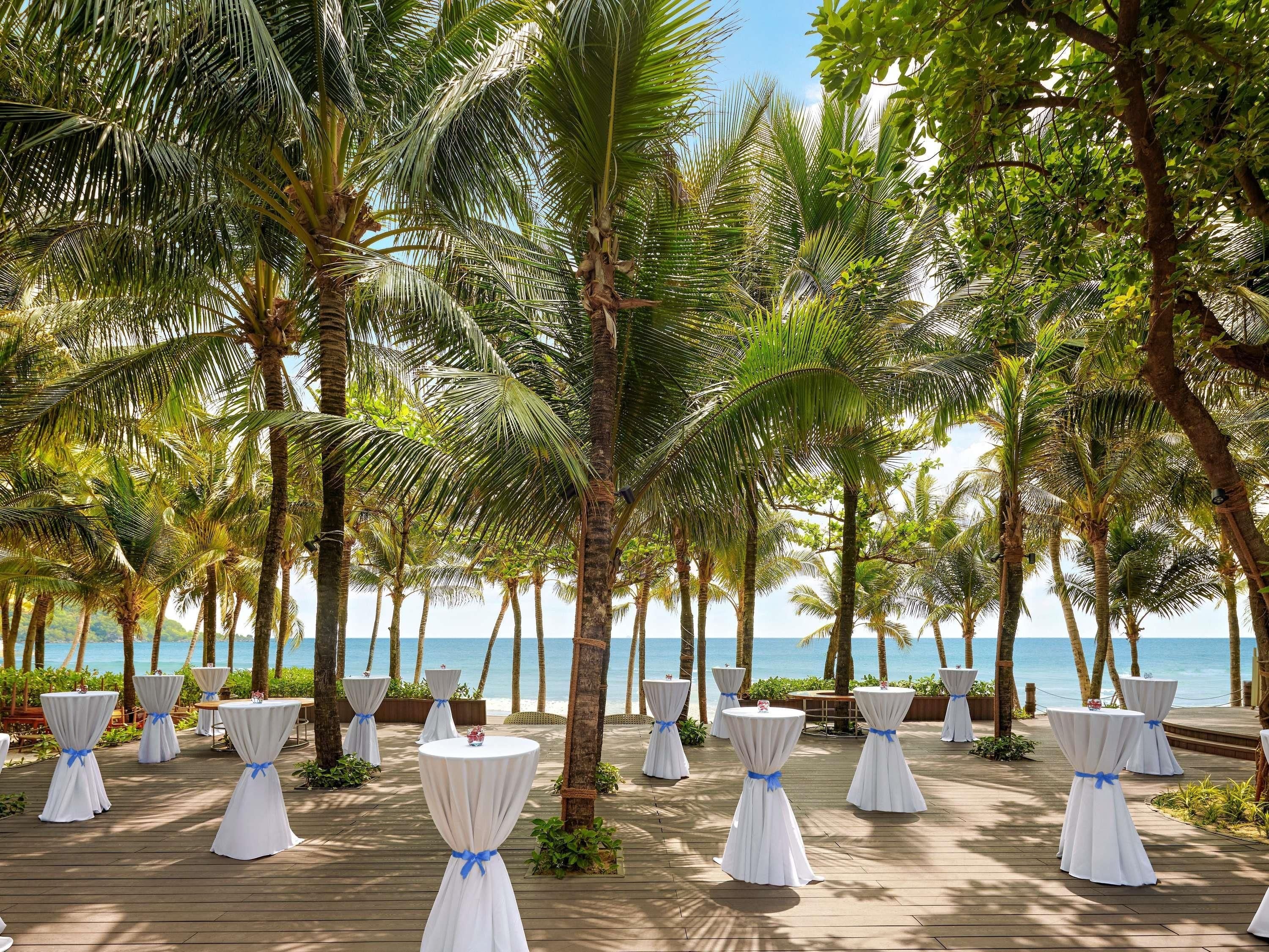A deck with round tables, each with a white tablecloth and a blue ribbon, surrounded by palm trees and overlooking a beach.