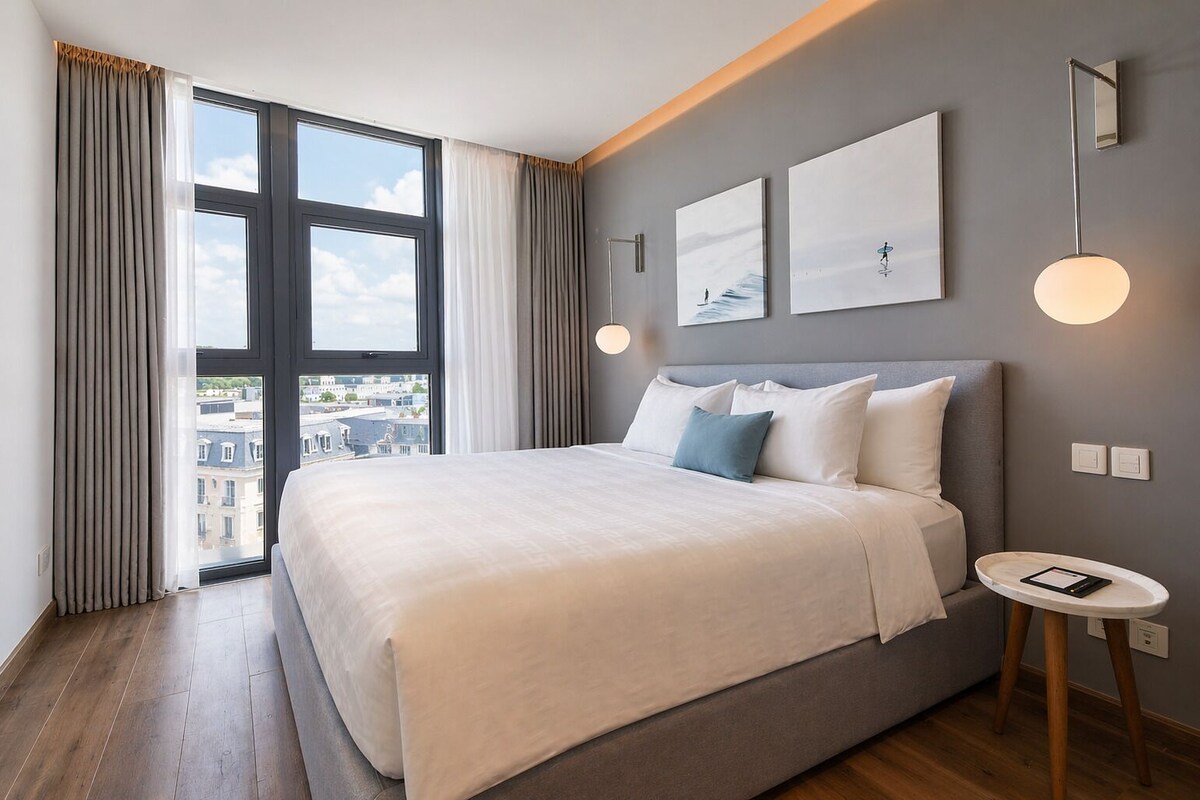 Una habitación de hotel moderna con una cama grande, una mesita de noche pequeña y vistas a la ciudad a través de amplias ventanas.