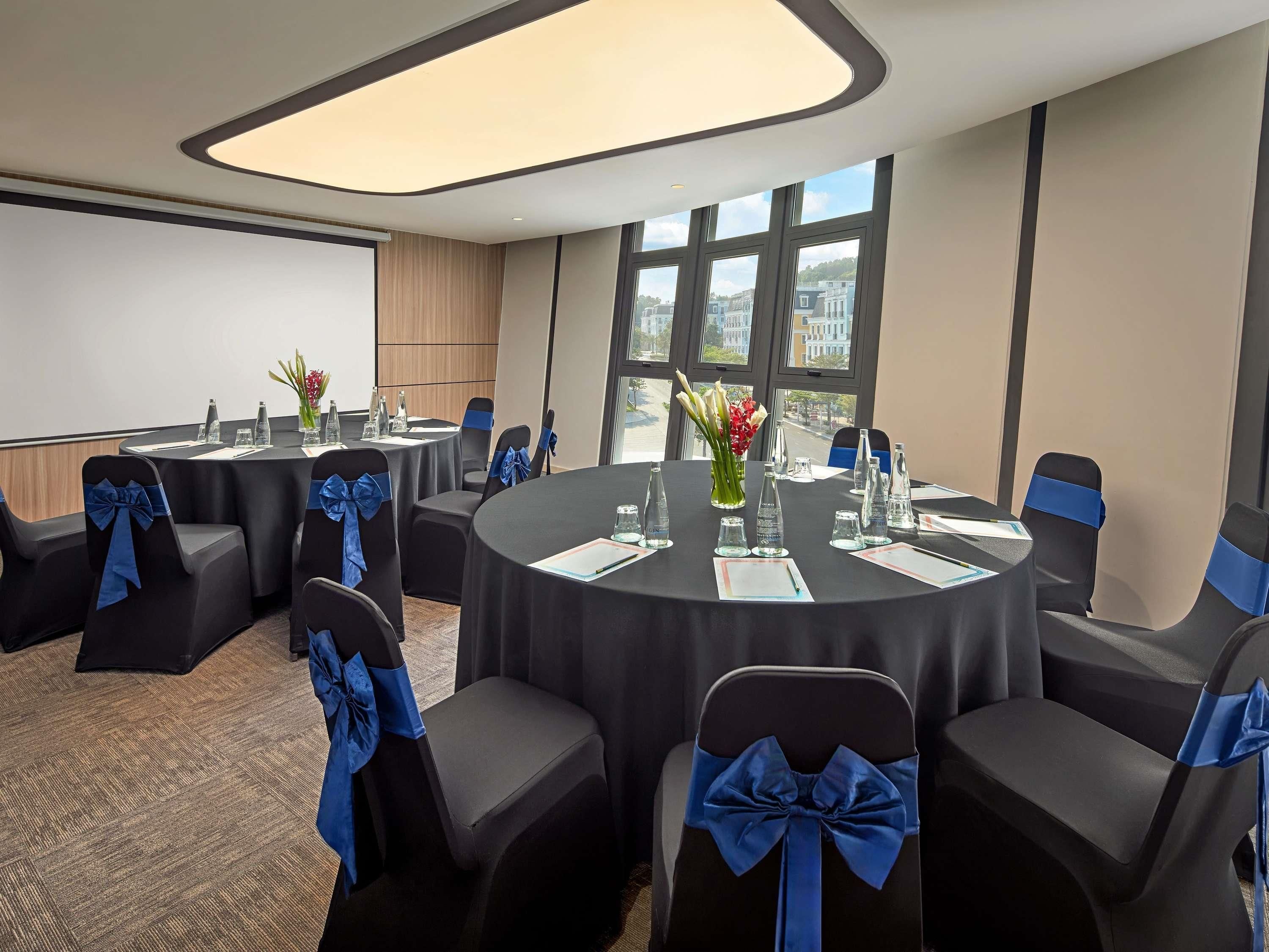 Une salle de conférence avec des tables rondes, des chaises ornées de rubans bleus et une grande fenêtre donnant sur des immeubles.