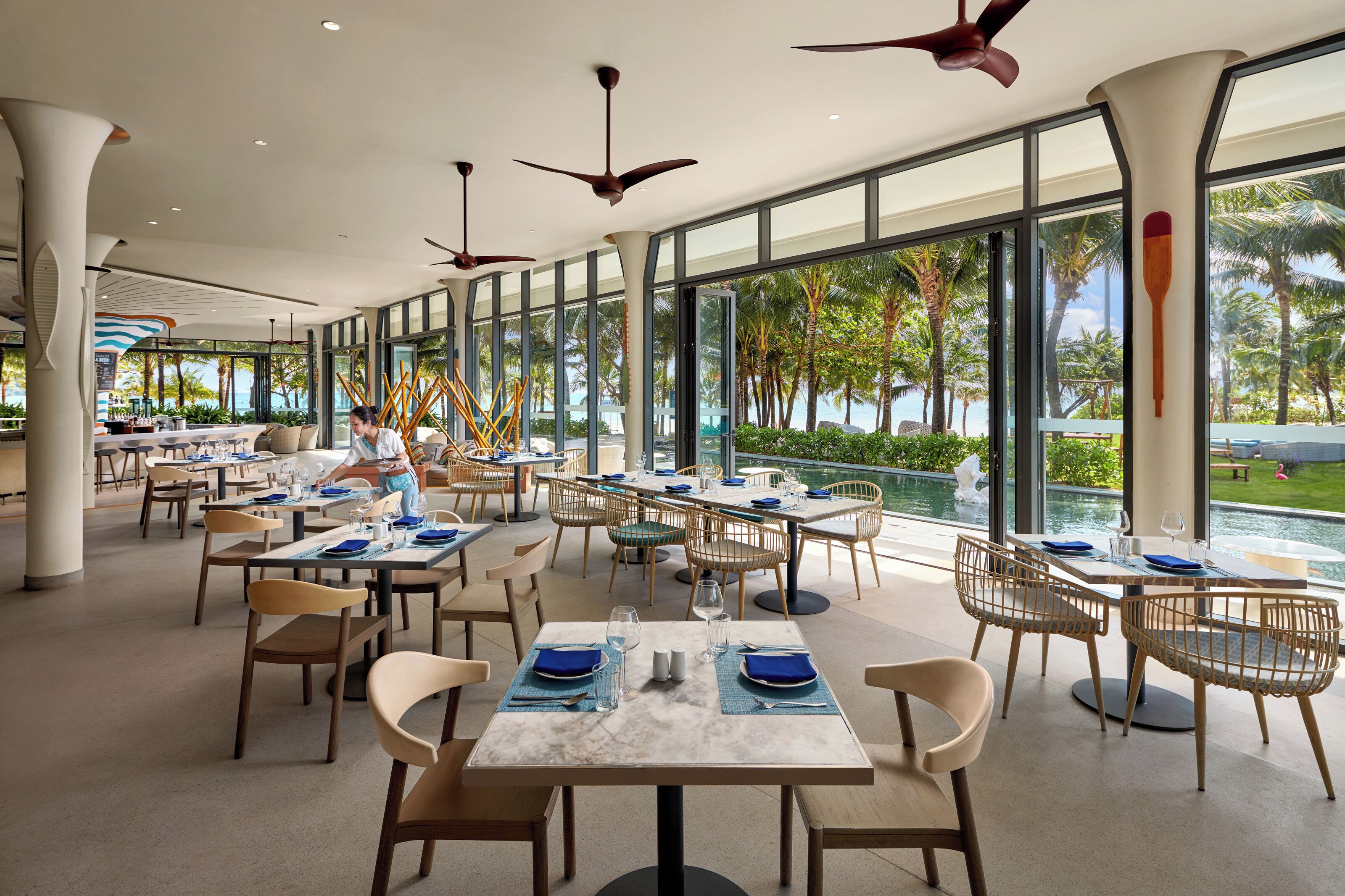 A spacious outdoor dining area with multiple tables and chairs, large windows offering views of palm trees and a pool.