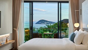 A hotel room with a large bed, a small table with chairs, and a view of a mountain and sea through floor-to-ceiling windows. - Premier Residences Phu Quoc Emerald Bay Managed by Accor (Phu Quoc)