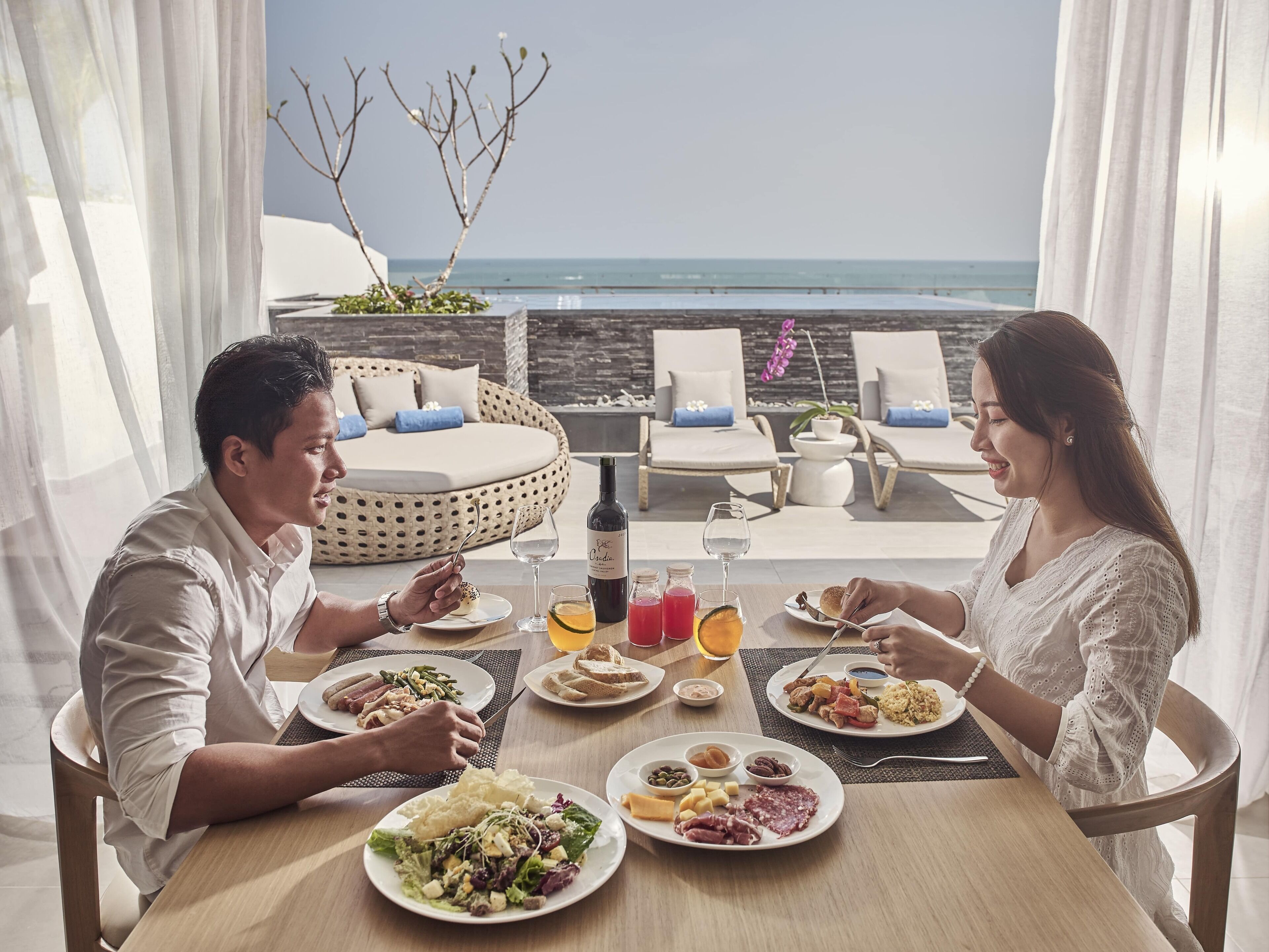 A dining table with plates of food, a bottle of wine, and a glass of orange juice. There are two people seated at the table. In the background, there is a bed with a view of the ocean and lounge chairs.