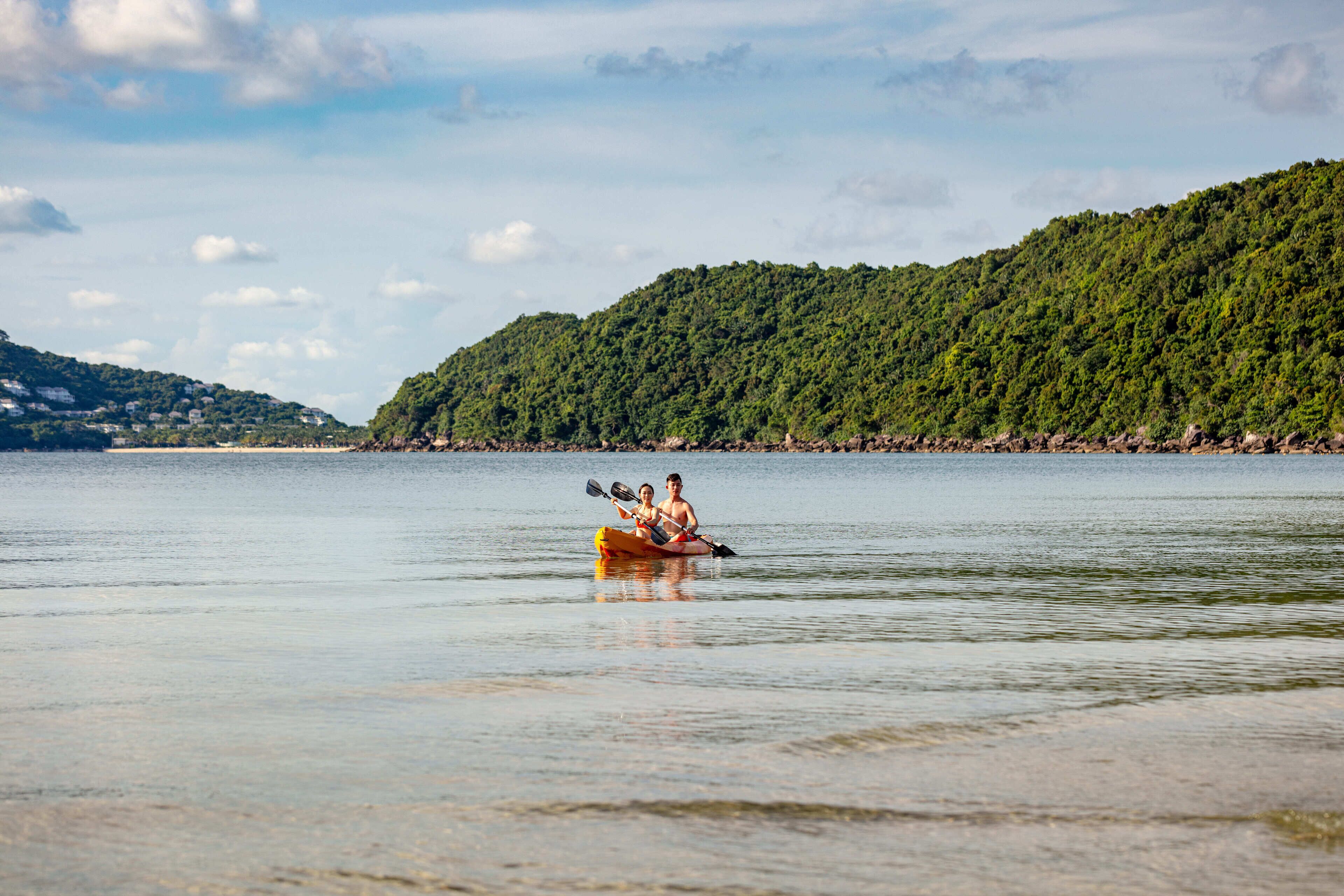 A couple kayaking on a calm lake with a forested hill in the background.