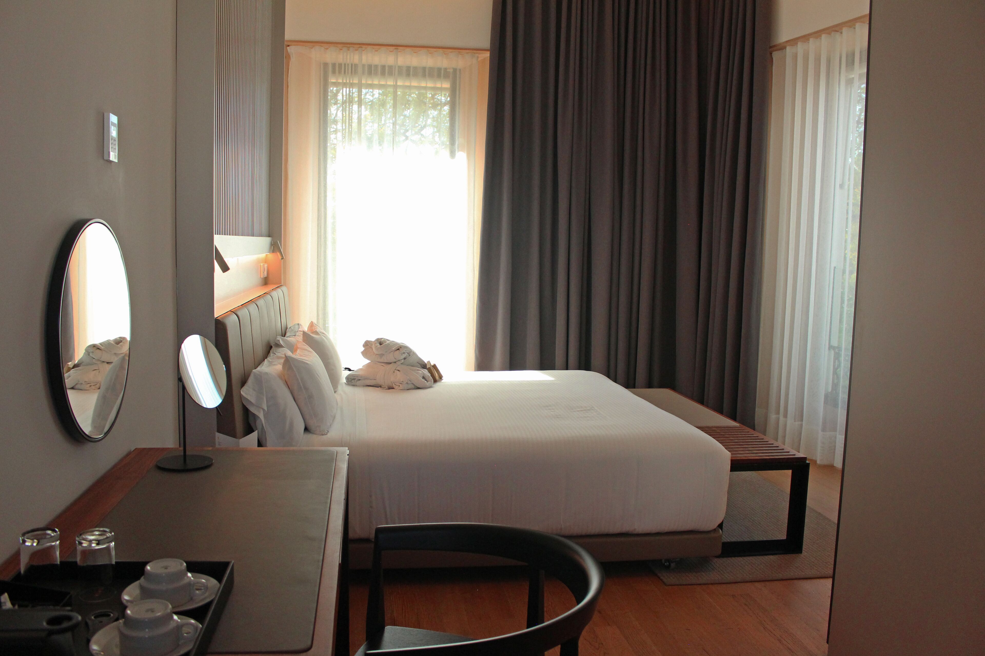 Habitación de hotel con cama, escritorio con espejo, silla y mesita de noche con una bandeja de tazas y un recipiente pequeño.