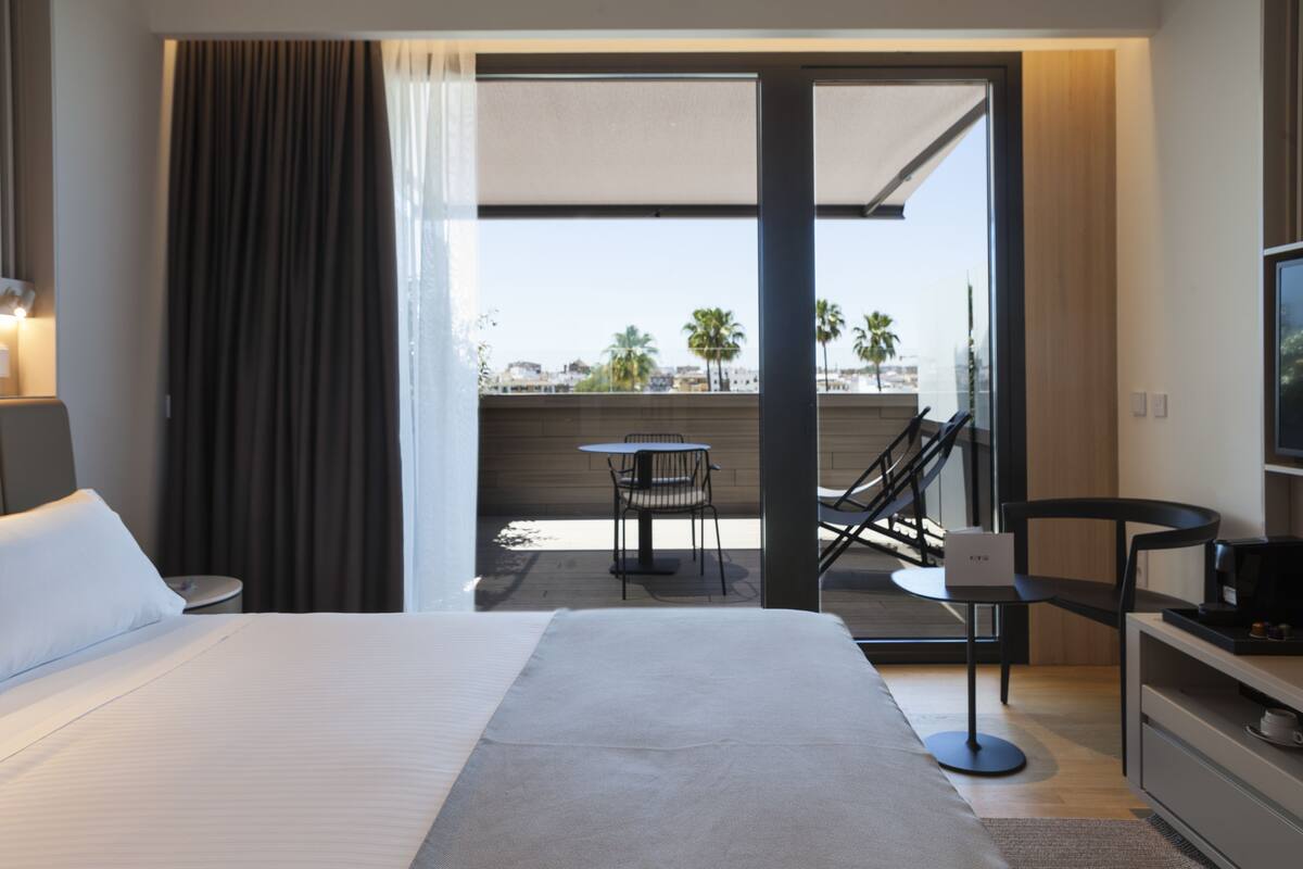 A modern hotel room with a large bed, a balcony with a table and chairs, and a TV on a stand.