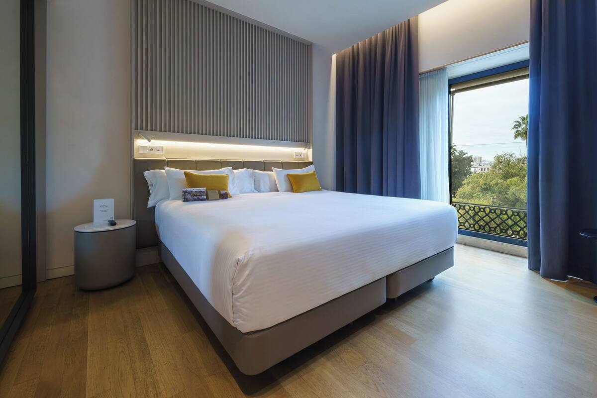 A modern hotel room with a large bed, a bedside table, a wall-mounted light, and a view of the outdoors.