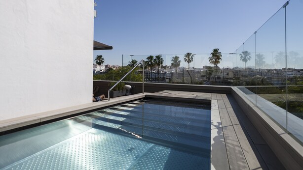 A rooftop pool with clear glass walls, a wooden deck, and a view of palm trees and buildings in the background. - Hotel Kivir Adults Recommended, Historic City Center (Seville)