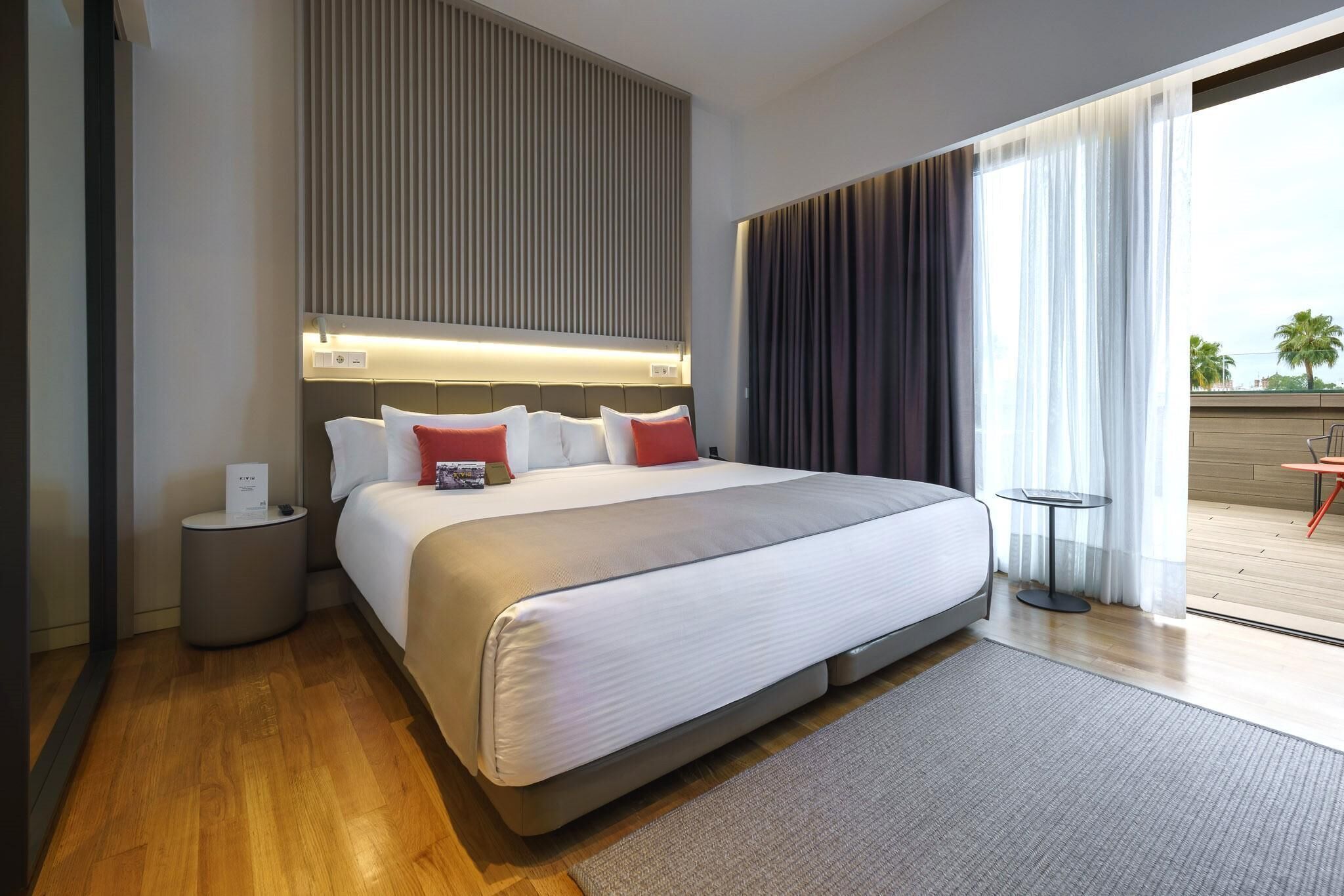 A modern hotel room with a large bed, a bedside table, a small round table, and a view of the outdoors.