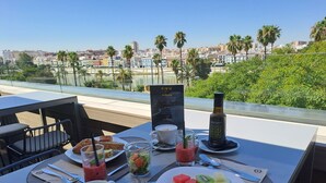 A table set with breakfast items including a plate of fruit, a glass of smoothie, a cup of coffee, and a bottle of water, with a city view in the background. - Hotel Kivir Adults Recommended, Historic City Center (Seville)