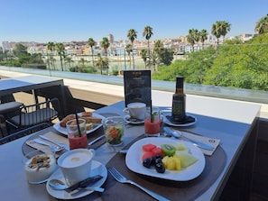 A table set with breakfast items including a plate of fruit, a glass of smoothie, a cup of coffee, and a bottle of water, with a city view in the background. - Hotel Kivir Adults Recommended, Historic City Center (Seville)
