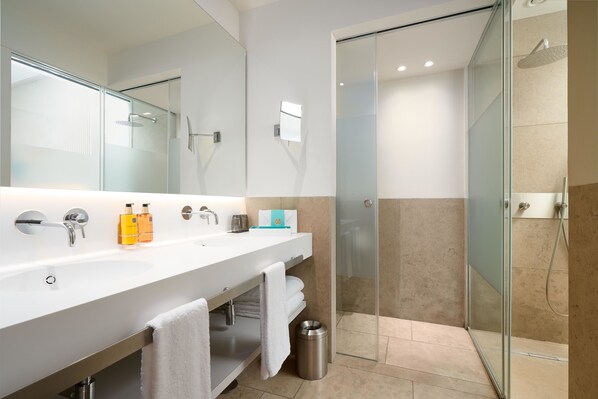 Un baño moderno con un espejo grande, dos lavamanos, una ducha con mampara de vidrio y un cesto de basura.
