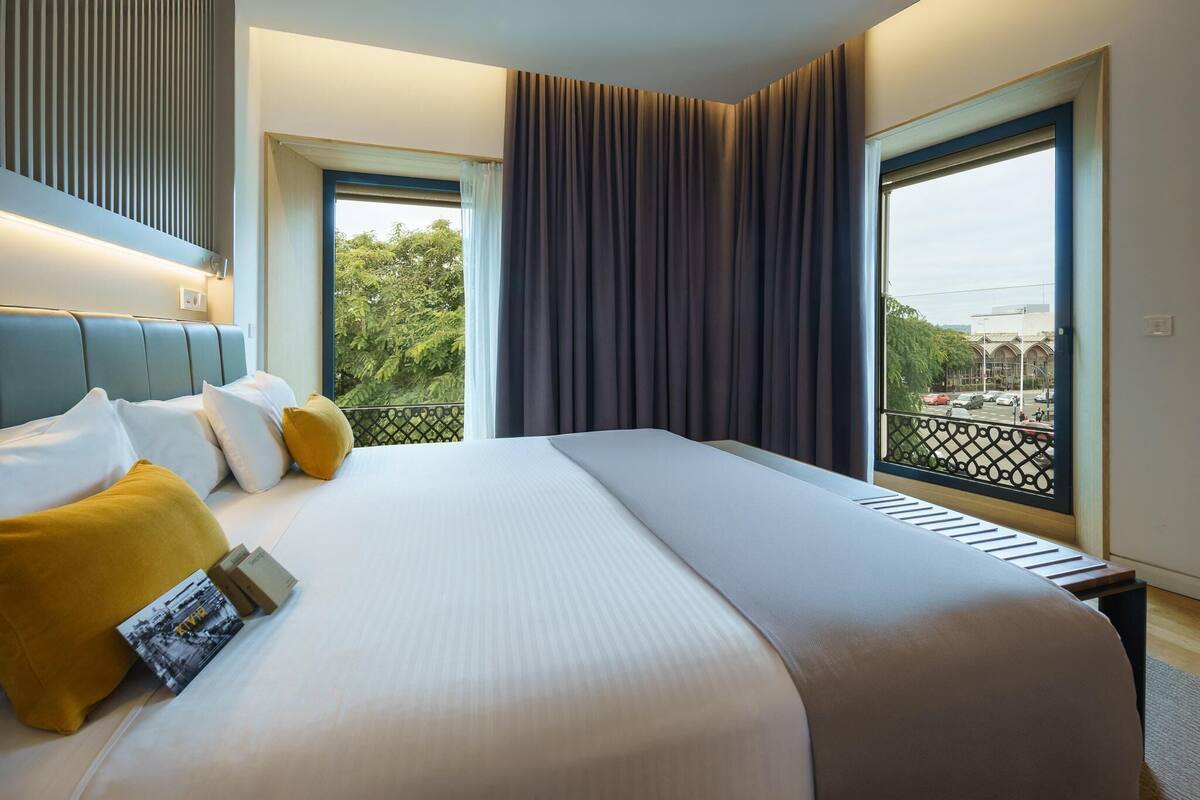 A hotel room with a large bed, a headboard, a window with a view of the city, and a balcony with a railing.