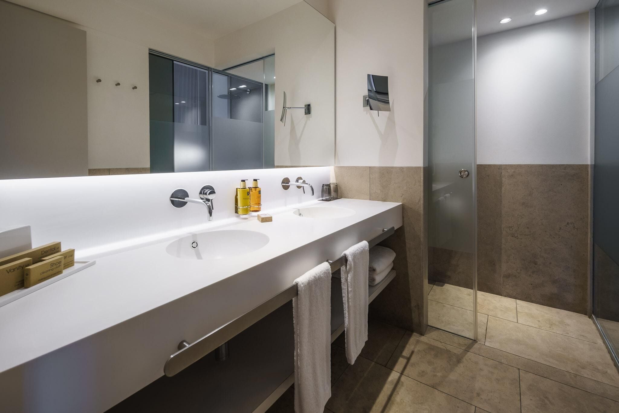Ein modernes Badezimmer mit einem langen, weißen Waschbecken, zwei Spiegeln und einer Duschkabine aus Glas.