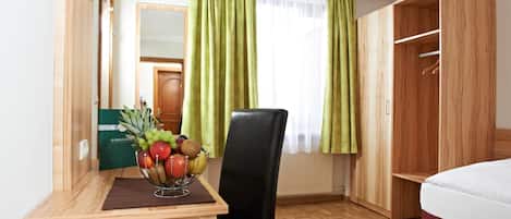 A hotel room with a wooden desk, a chair, a bed, a wardrobe, and a window with green curtains.