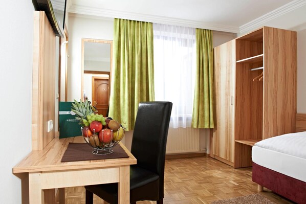 A hotel room with a wooden desk, a chair, a bed, a wardrobe, and a window with green curtains.