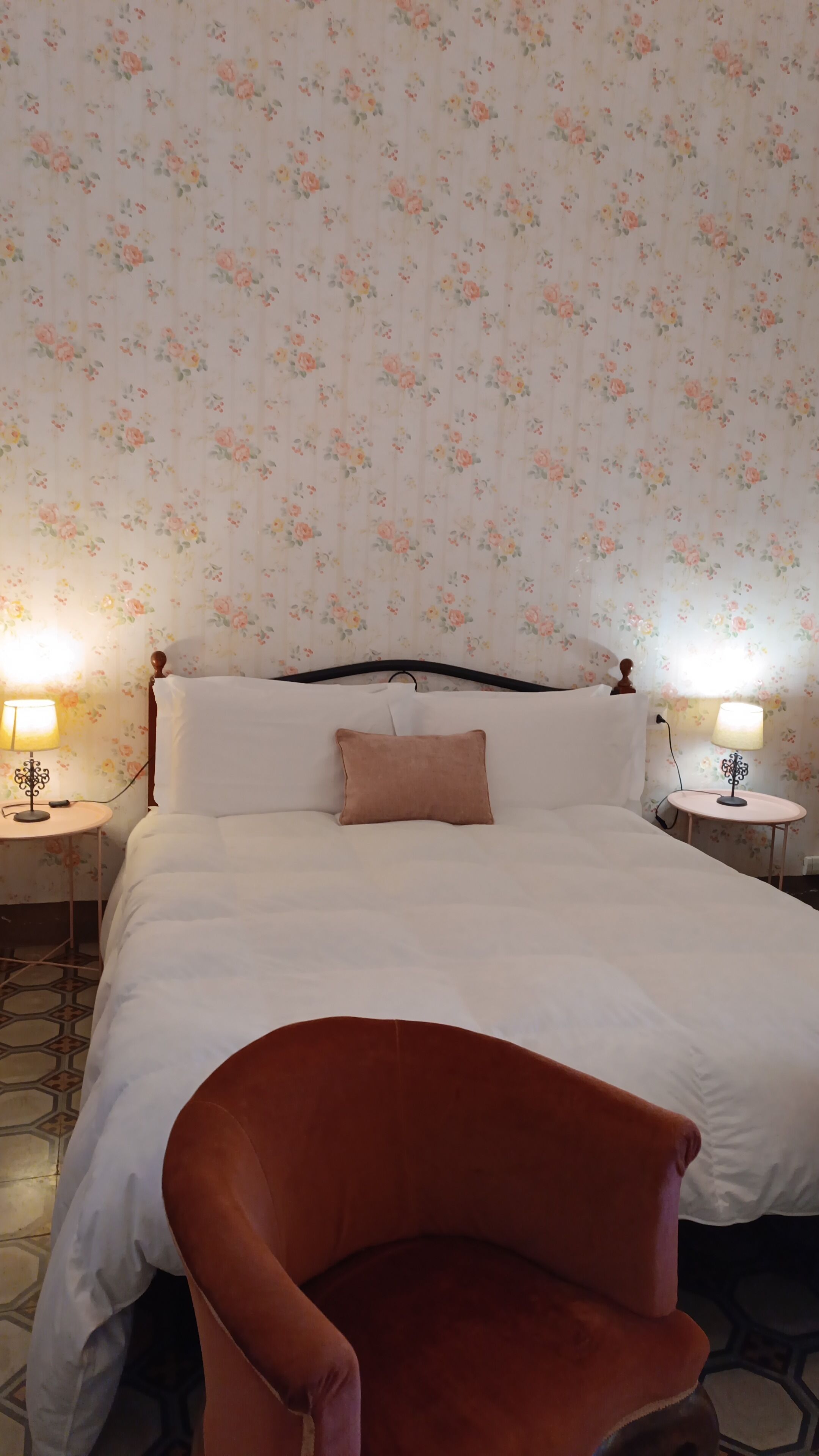 A neatly made bed with a headboard, two bedside lamps, and a floral wallpaper pattern.