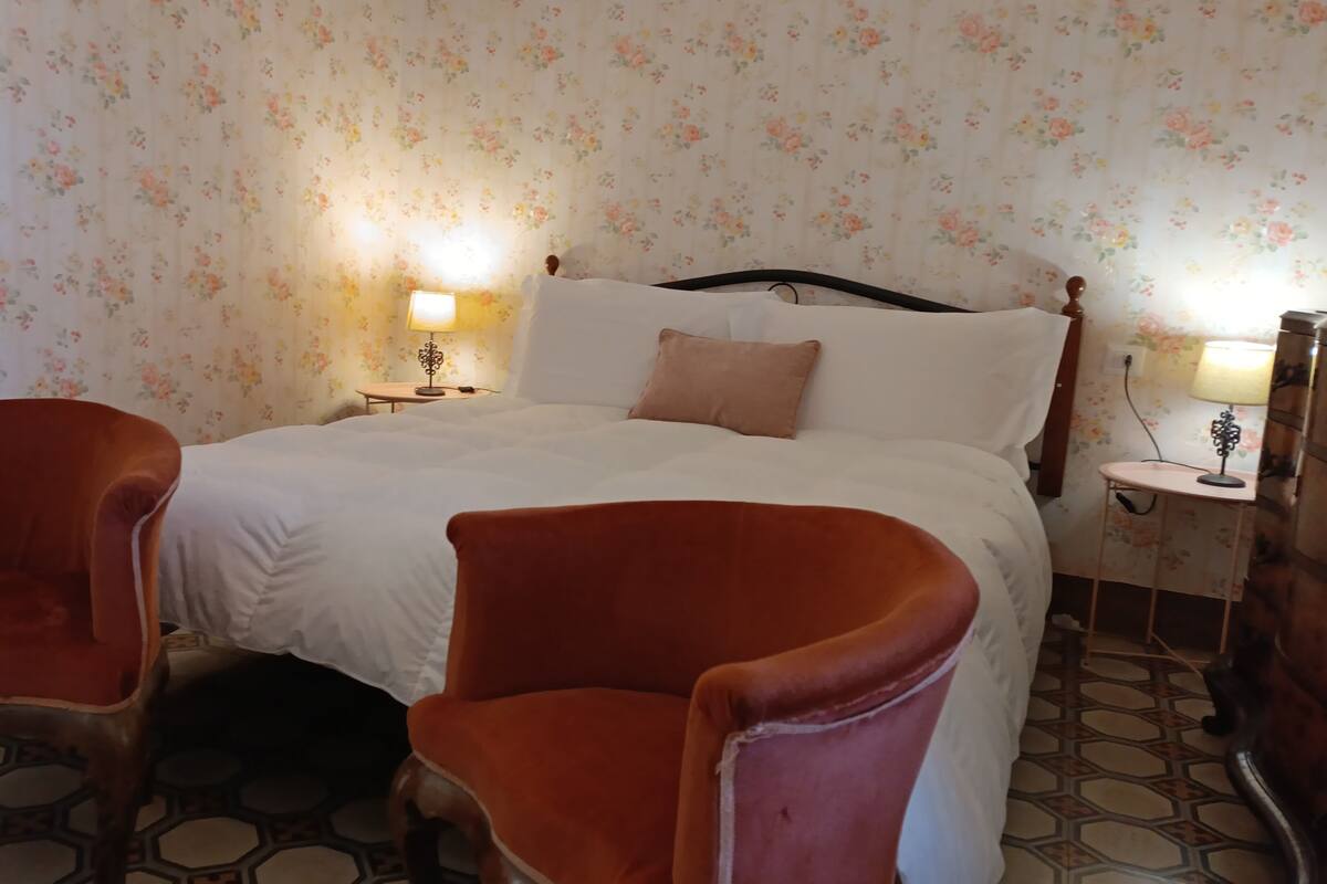 Ein Schlafzimmer mit einem Bett, zwei Sesseln, einem Nachttisch mit Lampe und blumigem Wandpapier.