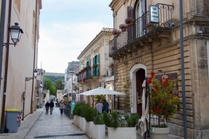 Une rue étroite bordée de bâtiments des deux côtés, avec des tables et des chaises installées à l’extérieur sous des parasols, et une enseigne indiquant un bed and breakfast.