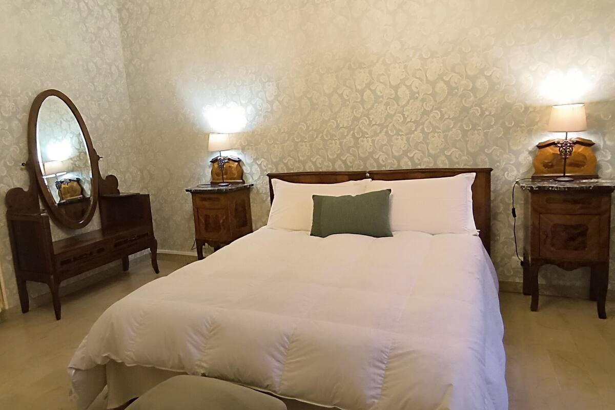 Ein ordentlich eingerichtetes Schlafzimmer mit einem großen Bett, zwei Nachttischen mit Lampen, einer Kommode und einem Spiegel.