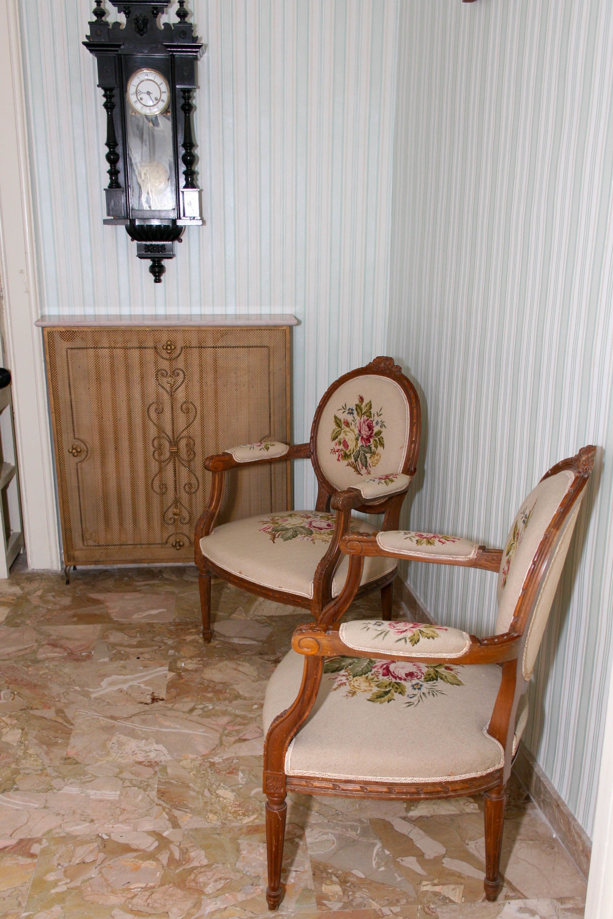 Deux chaises en bois recouvertes d’un tissu à motifs floraux, un meuble en bois et une horloge murale.