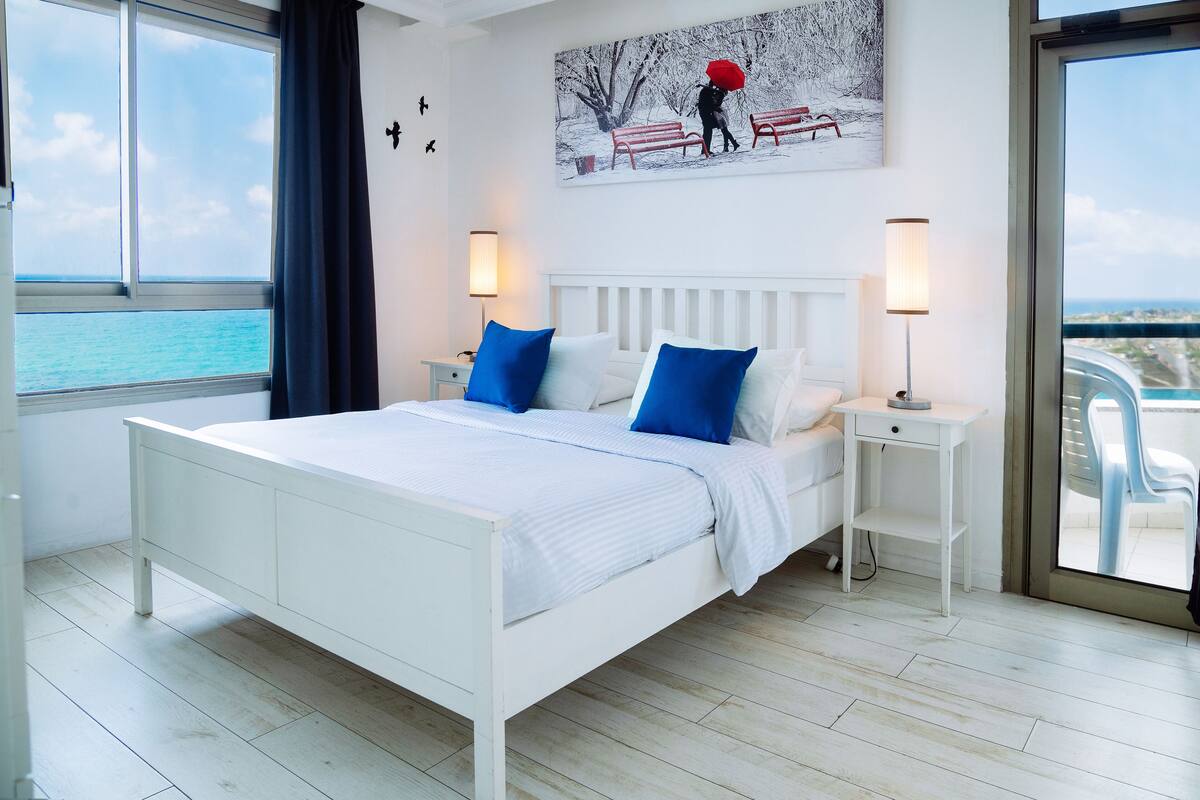 A modern bedroom with a large bed, two bedside tables, and a view of the sea.