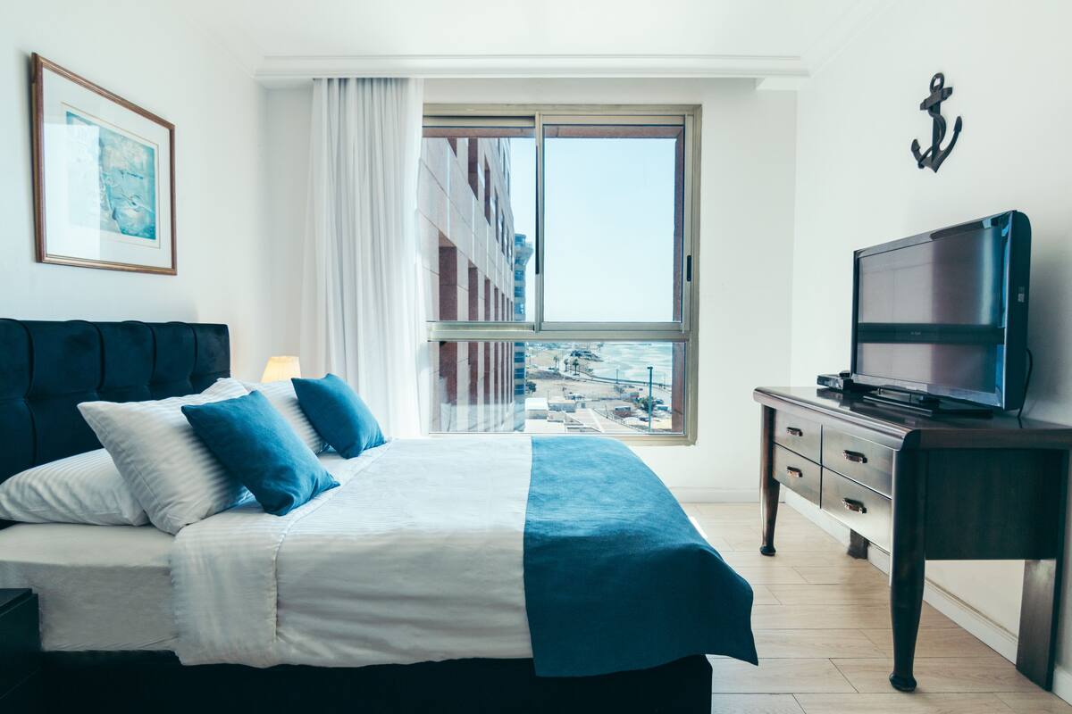 A hotel room with a bed, a TV, a window with a view of buildings, and a wall-mounted anchor decoration.