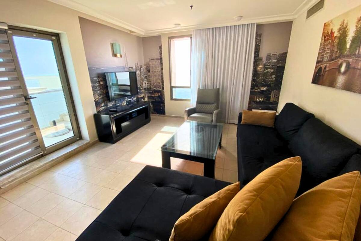 A living room with a black sofa, a glass coffee table, and a TV on a black stand. There is a cityscape mural on the wall and a window with curtains.