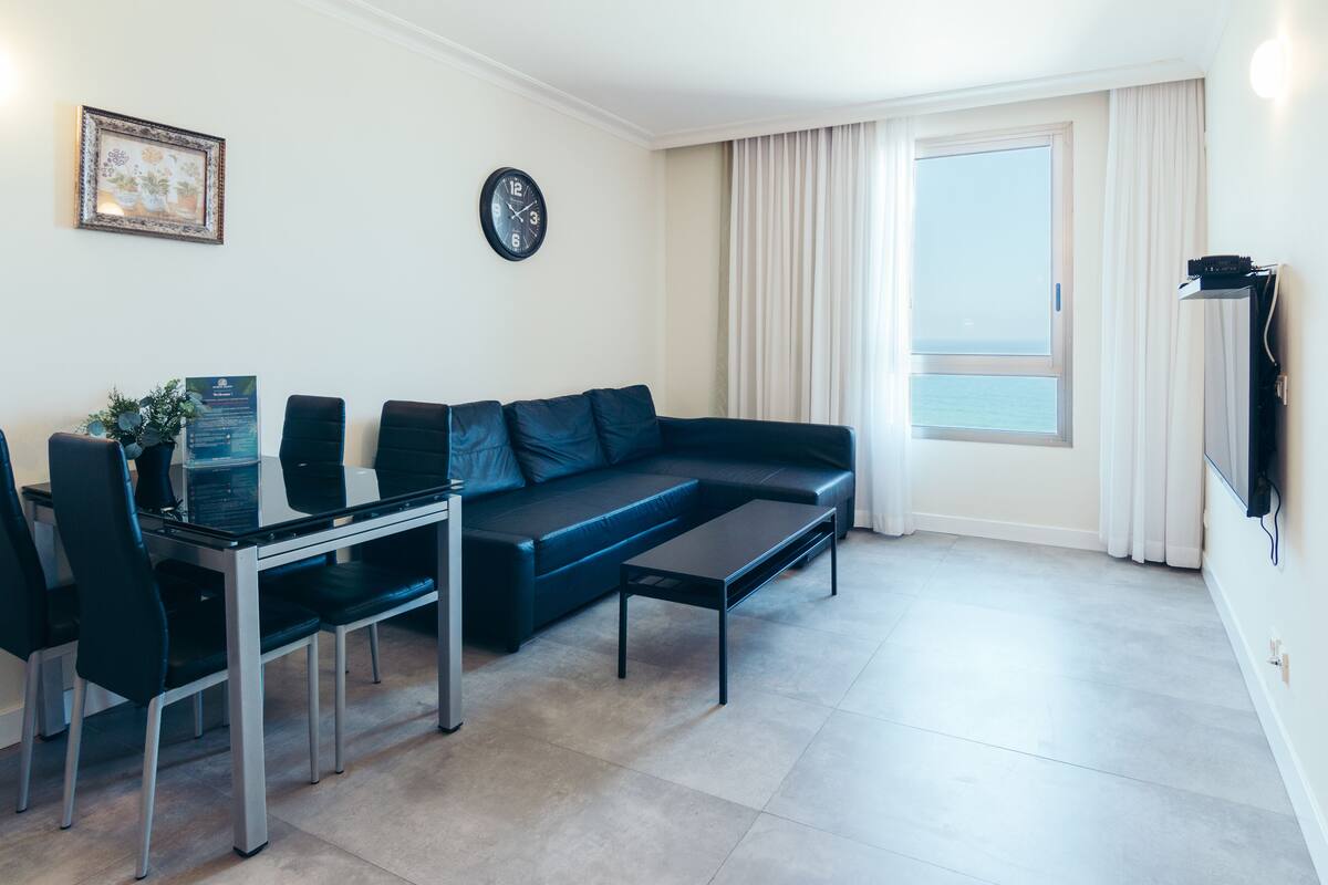 A modern living room with a sectional sofa, a glass coffee table, and a dining area with chairs and a table. A clock and a framed picture are on the wall.