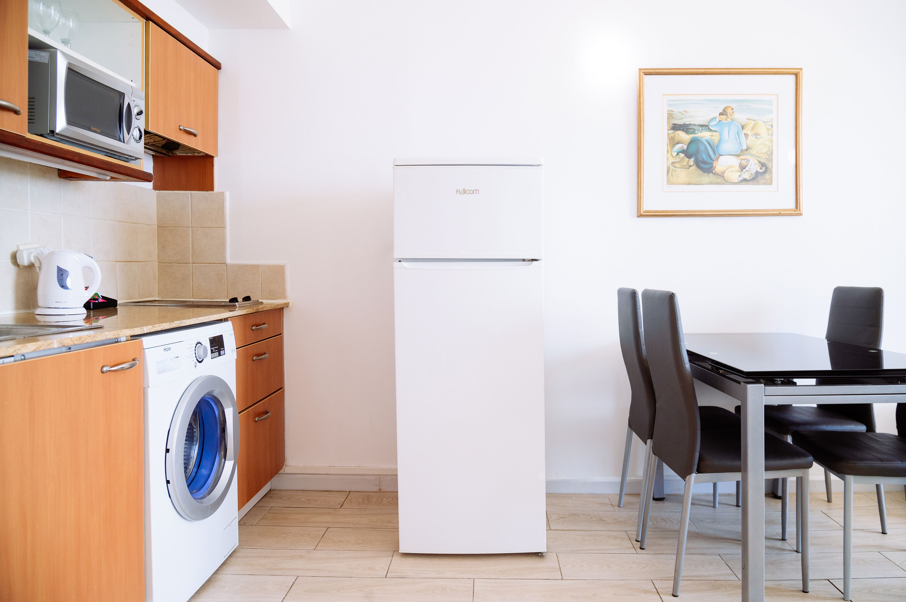 A compact kitchen with a washing machine, a microwave, and a refrigerator. There is a dining table with chairs and a framed picture on the wall.