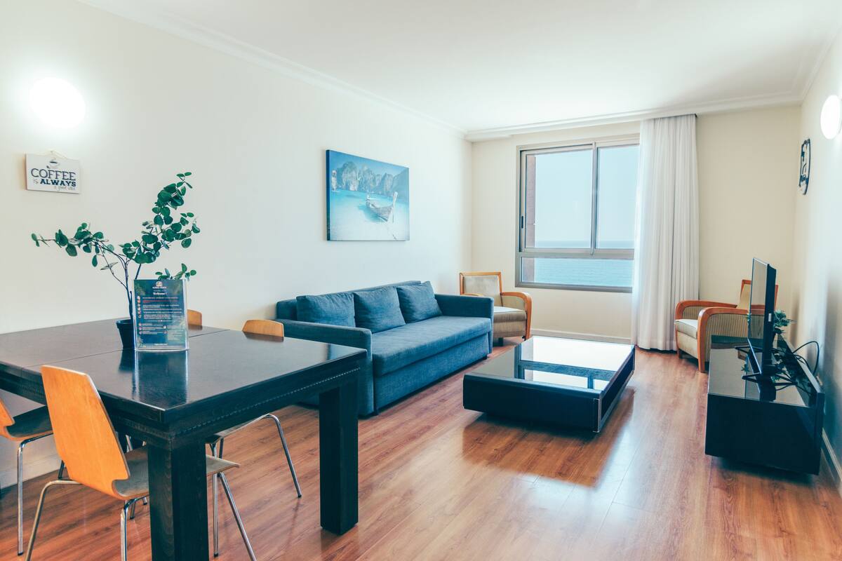 A modern living room with a dark wood floor, a blue sofa, a coffee table, and a dining table with chairs. There is a large window with curtains, a painting on the wall, and a sign that says "Coffee is Always".