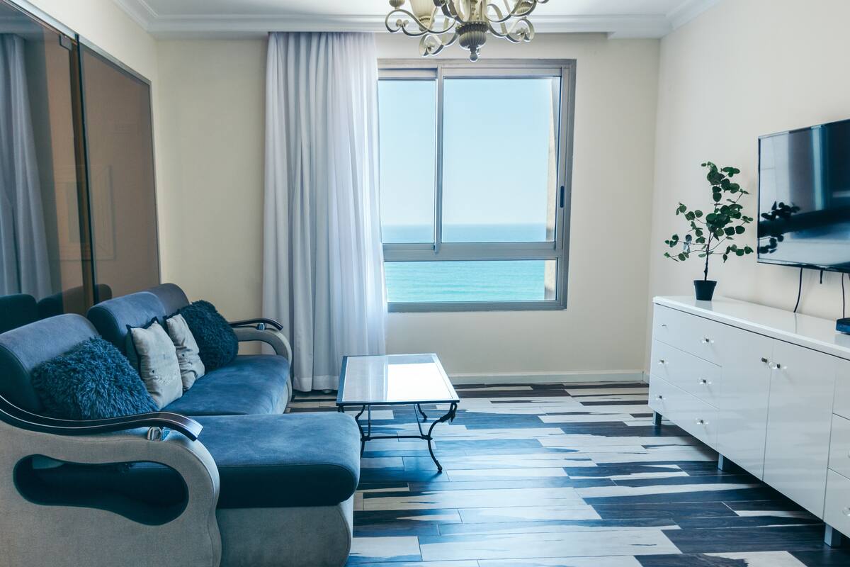 A modern living room with a sofa, a coffee table, a TV, and a view of the sea through a large window.