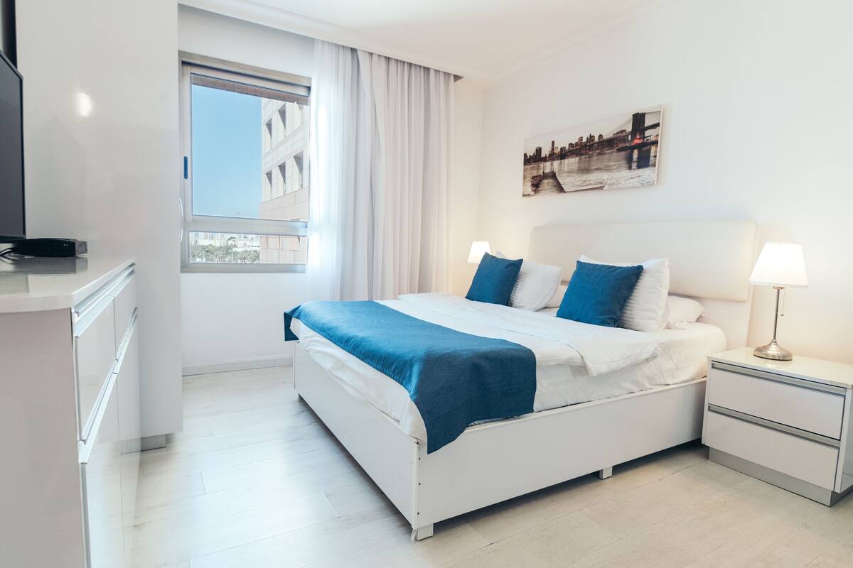 A modern hotel room with a bed, bedside tables, a TV, and a window with curtains.