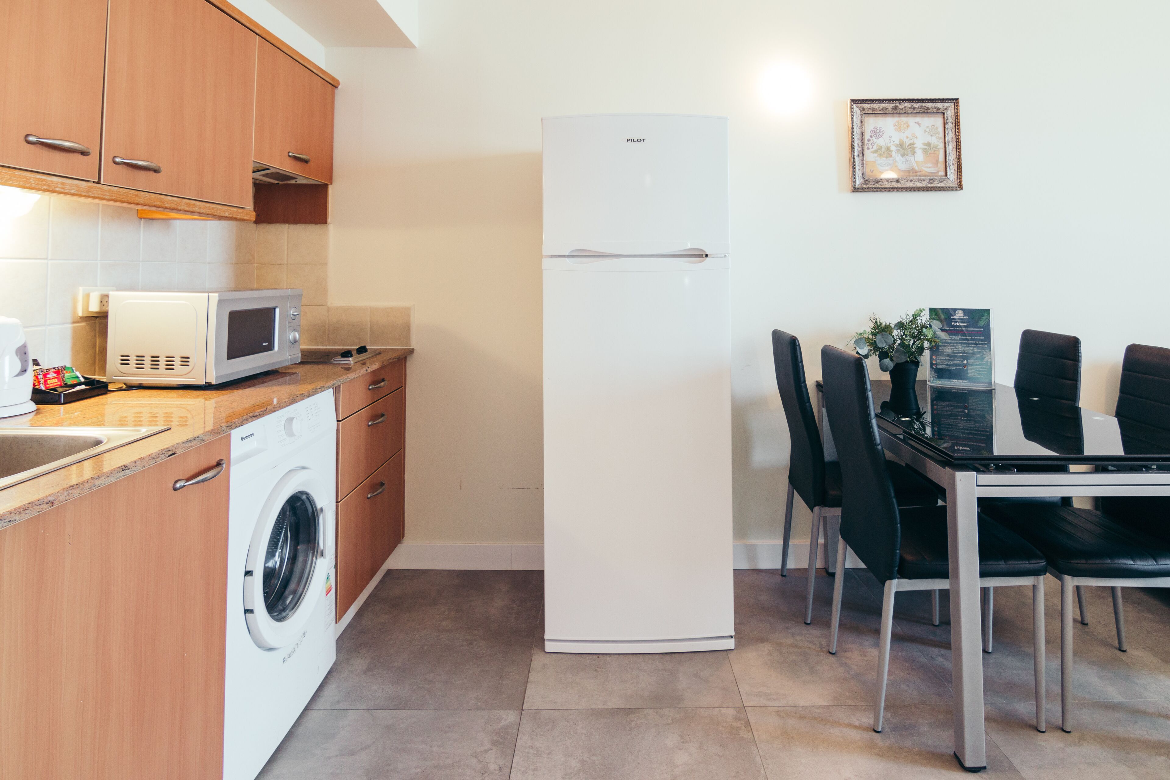 Una cocina funcional con armarios de madera, lavadora, microondas, refrigerador blanco y una mesa de comedor con sillas.