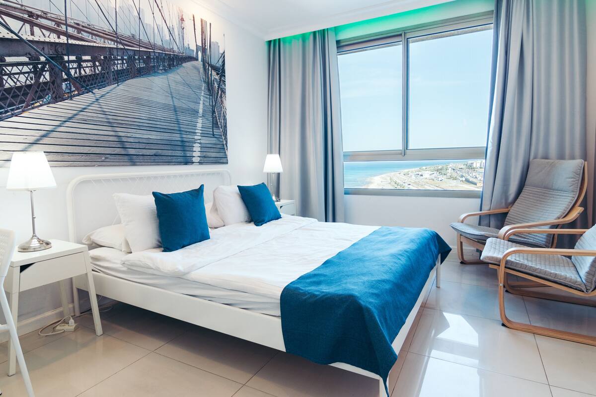 A modern hotel room with a bed, a chair, a desk, and a large window with a view of the beach.